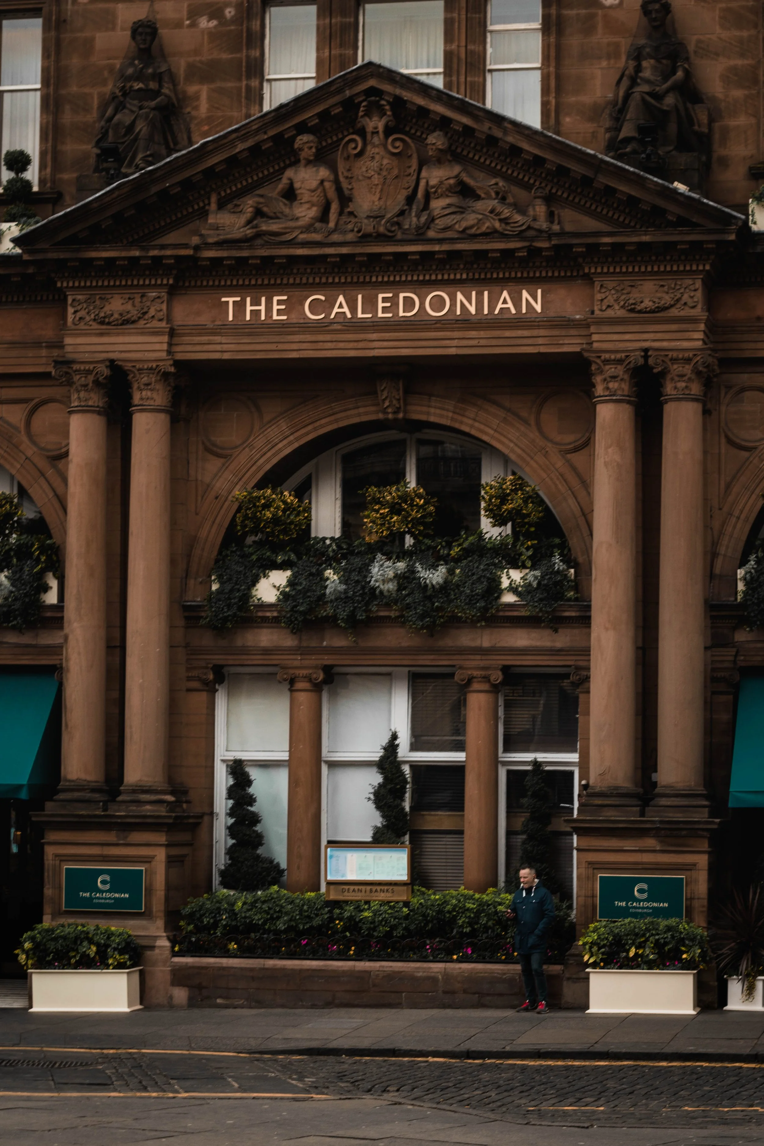 The exterior of the Caledonian building, featuring classical architecture with columns, sculptures, and greenery, with a man standing on the sidewalk.