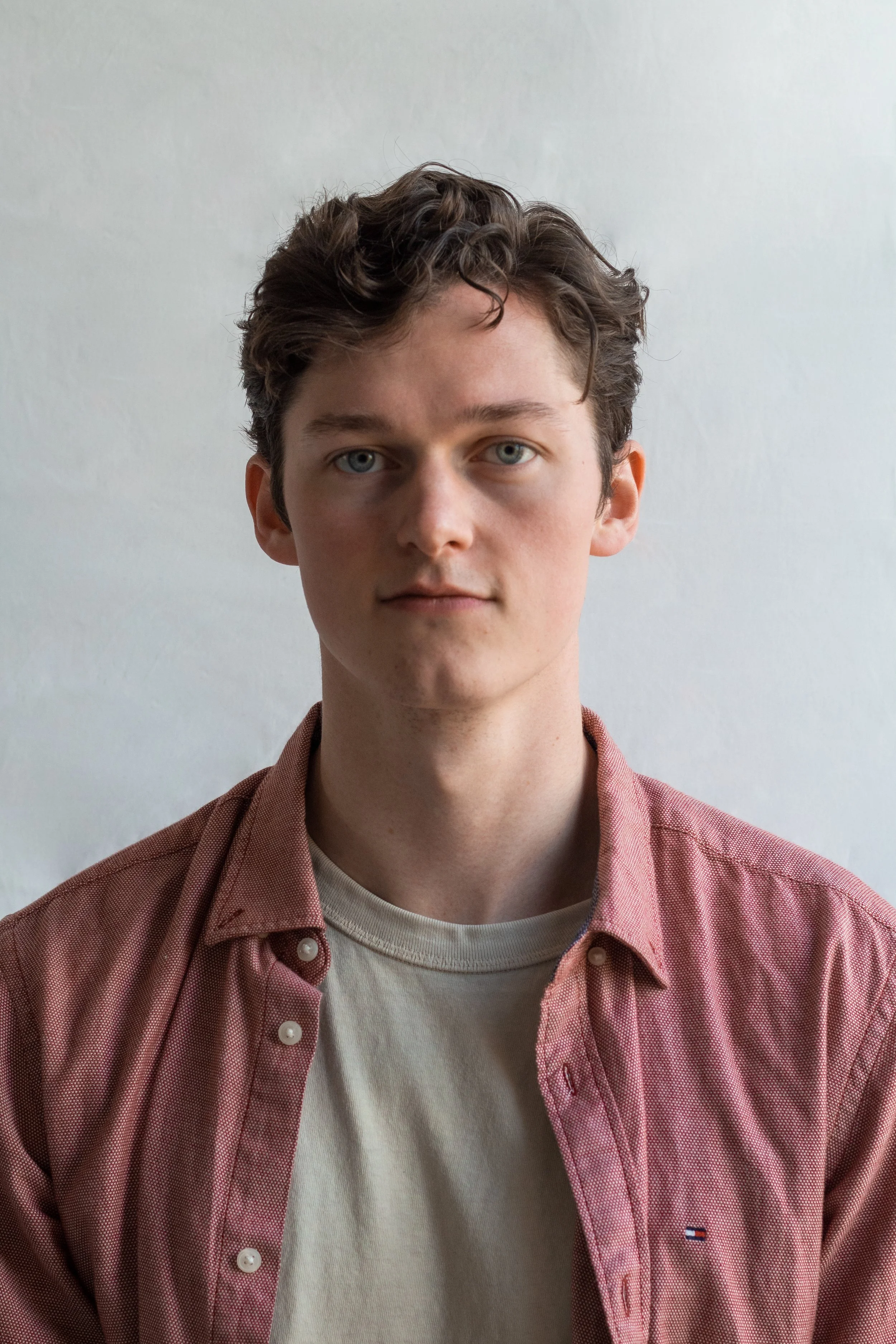 A young man with wavy brown hair, blue eyes, and fair skin, wearing a light beige T-shirt and pinkish button-up shirt with a small logo on the chest, standing against a plain white background.