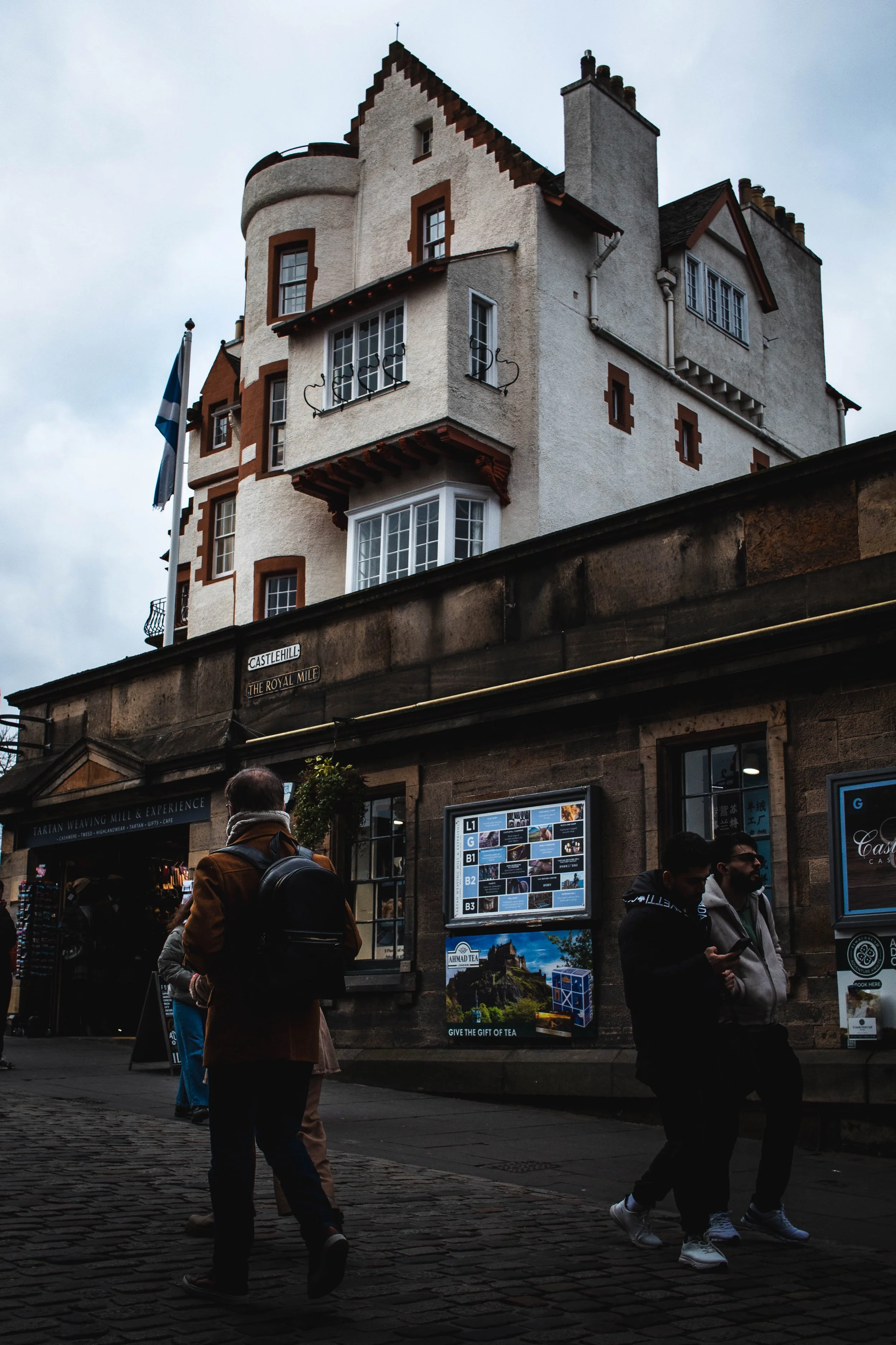 A tall, historic stone building with a white exterior, multiple windows, and a small balcony, situated on a cobblestone street with a few pedestrians walking by.