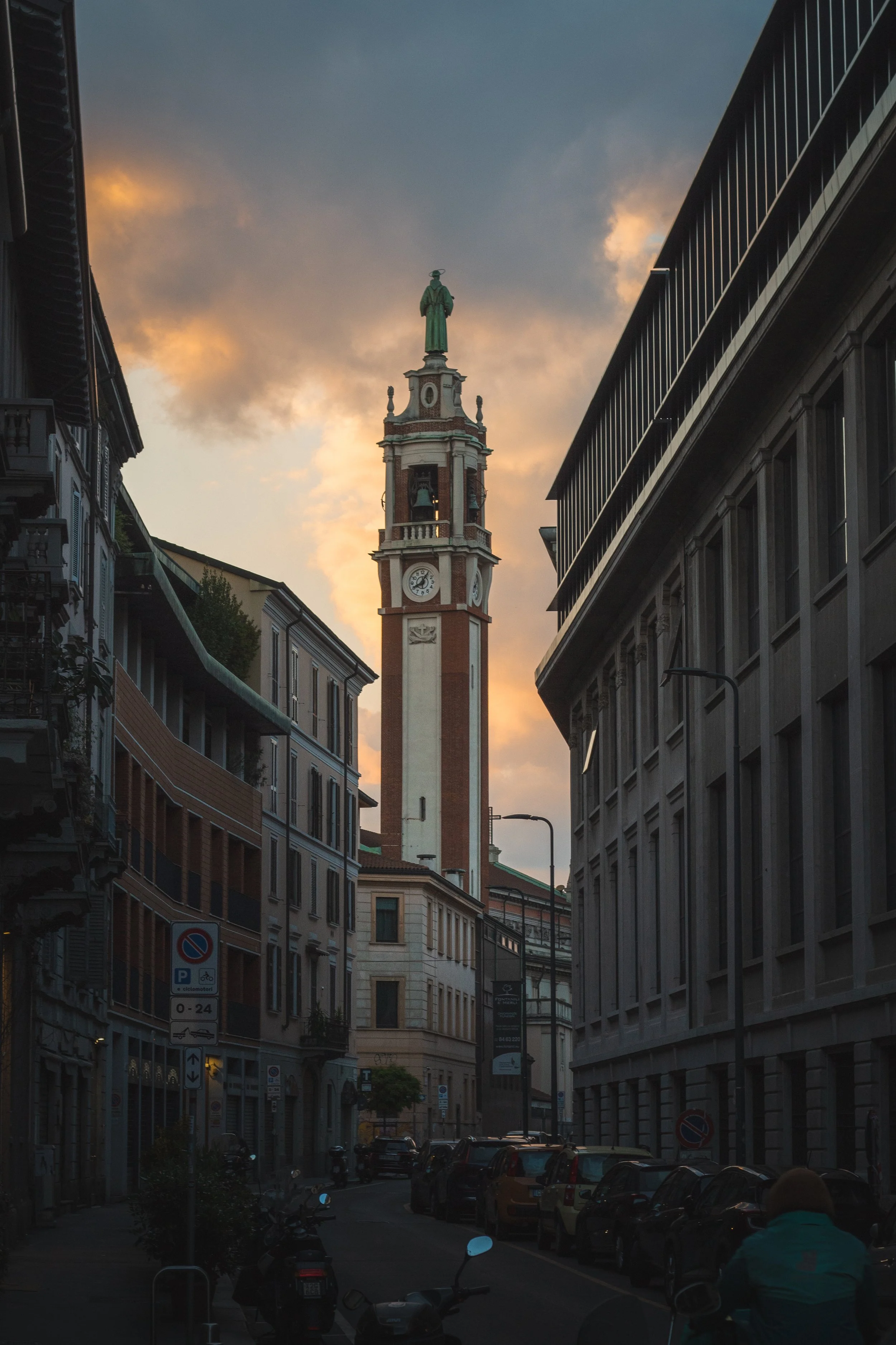 A city street at dusk with a tall clock tower topped by a statue in the background, surrounded by mid-rise buildings, parked cars, and street lamps.