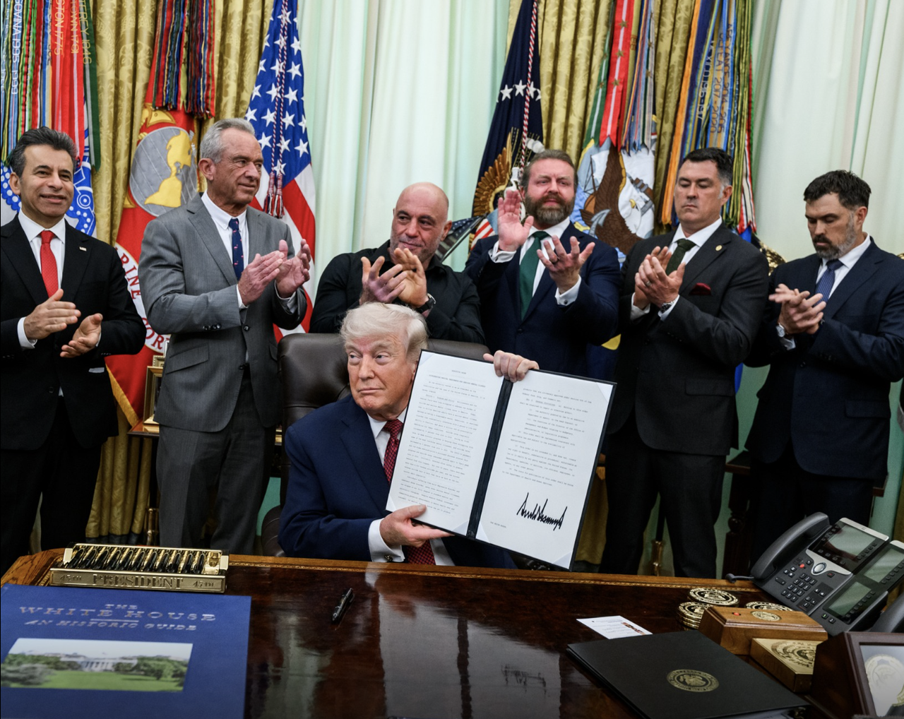 President Donald Trump holds up a signed executive order. He is flanked by a group of clapping men.