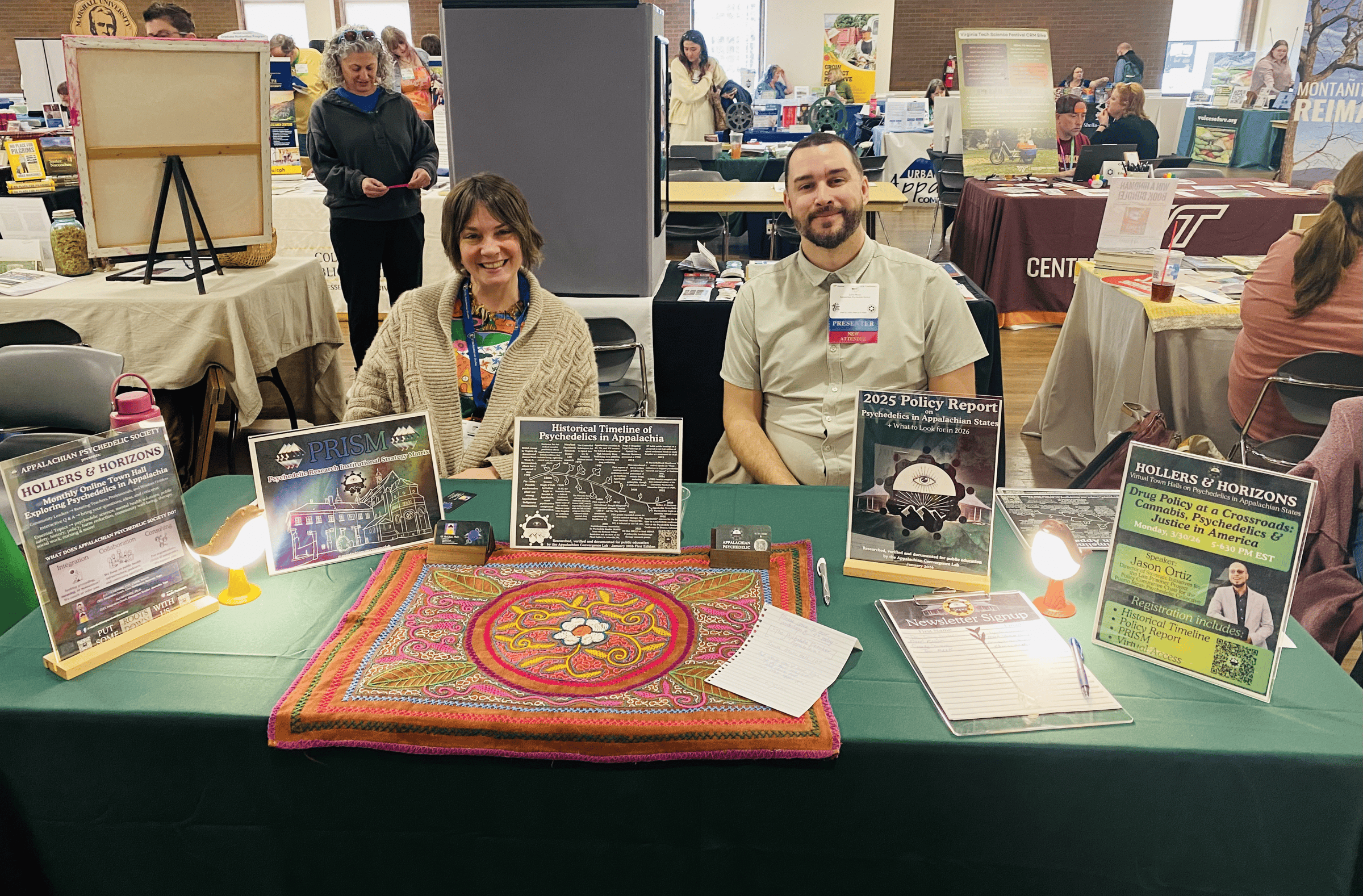 Two smiling people sit at a booth displaying flyers and a woven textile.