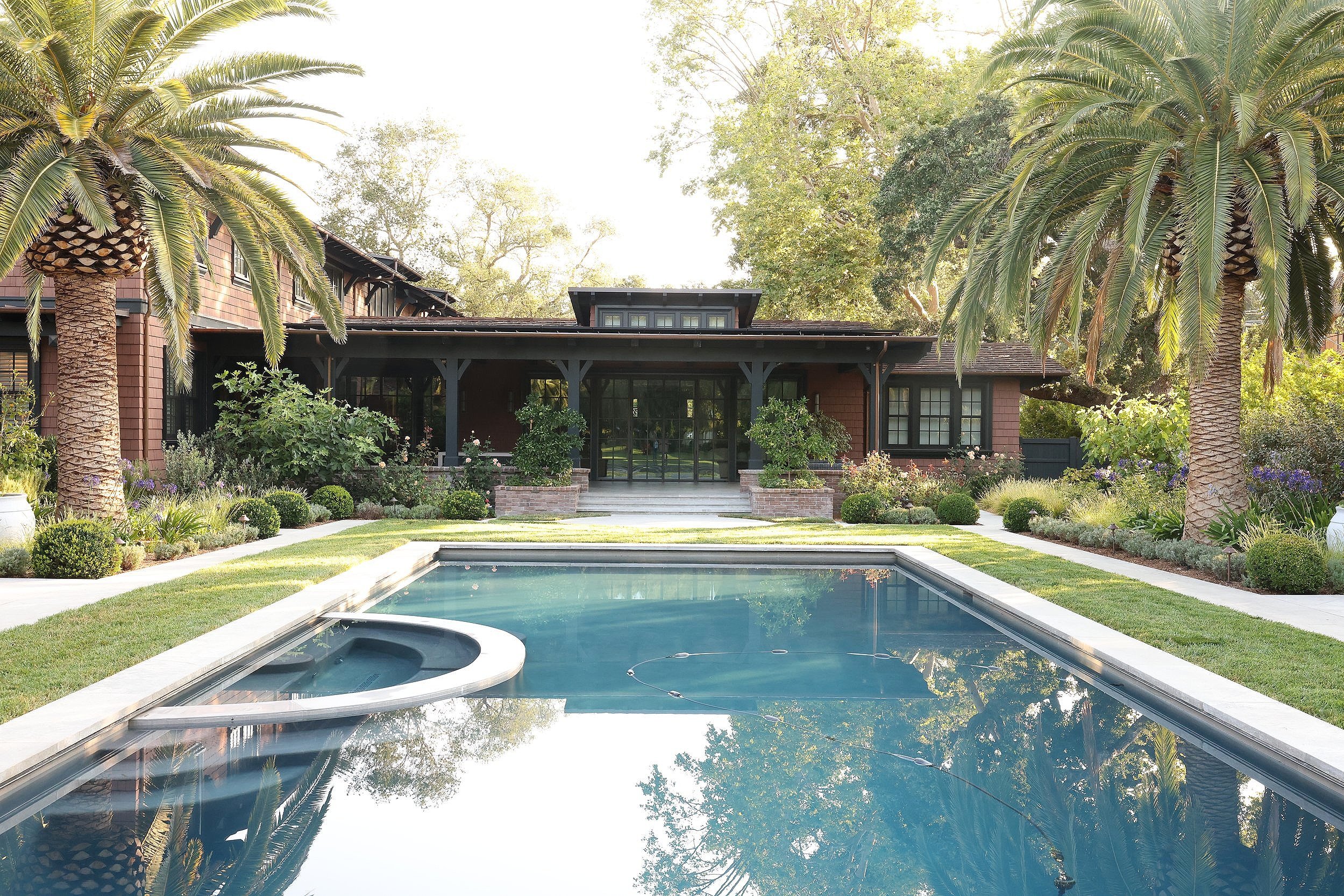 A backyard with a swimming pool, surrounded by grass, palm trees, and bushes, with a house in the background and sunlight filtering through the trees.