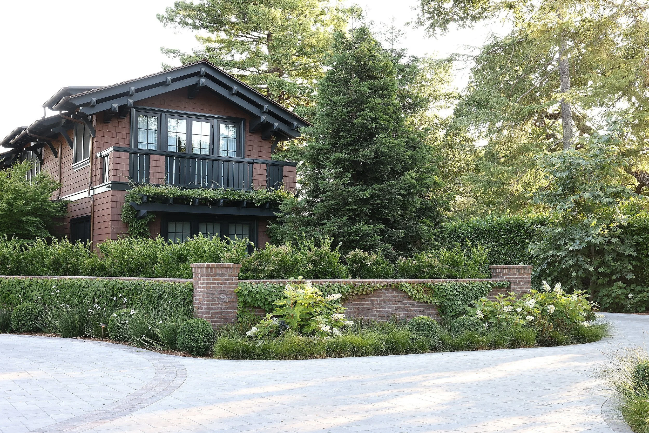 A two-story house with a balcony, surrounded by lush green trees and bushes. The house has wooden shingles and black window frames. A brick wall and neatly landscaped garden are visible in the foreground.