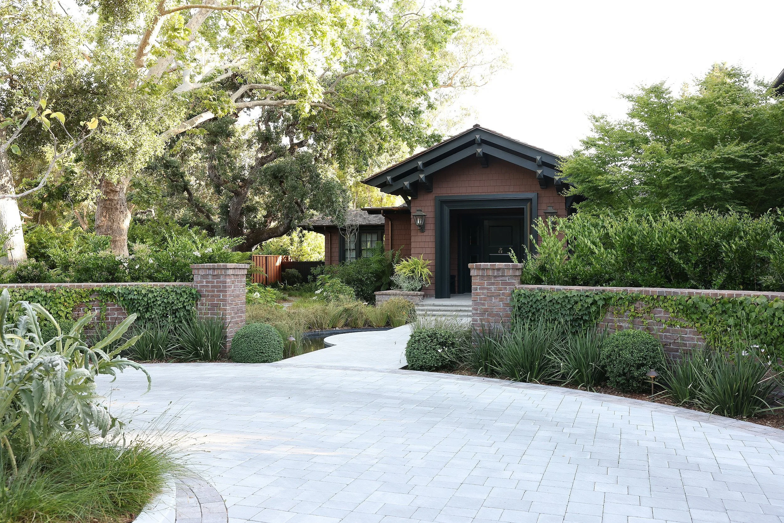 A house surrounded by lush greenery and trees, with a white paved pathway leading to the entrance.