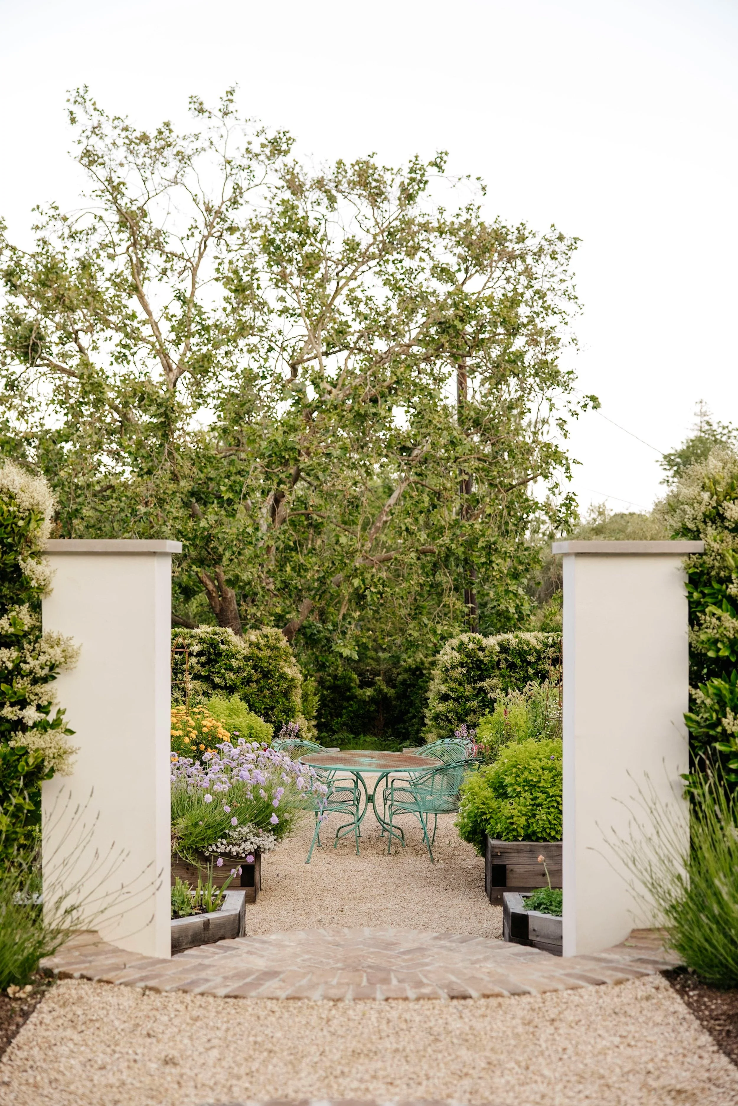 View of a lush garden with a round metal table and chairs, surrounded by vibrant flowers and greenery, framed by white gate posts.