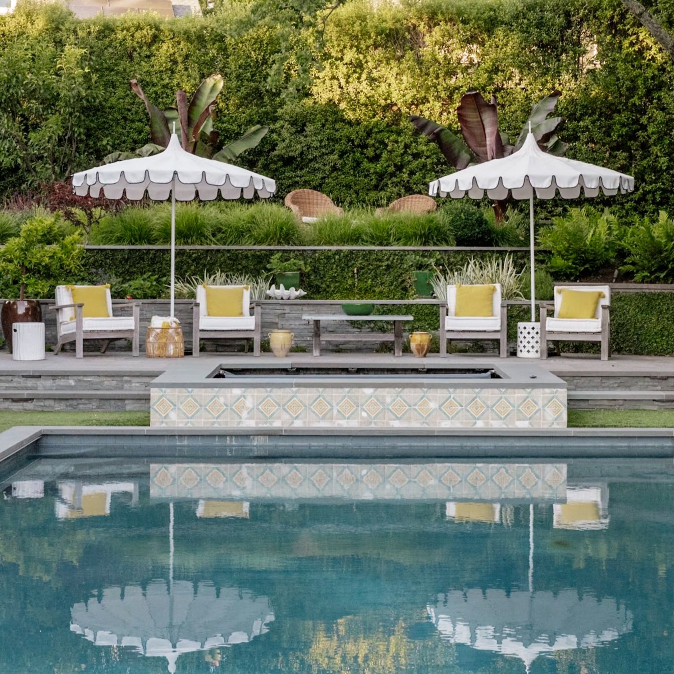 A backyard pool area with lounge chairs, large white umbrellas, potted plants, and greenery in the background.