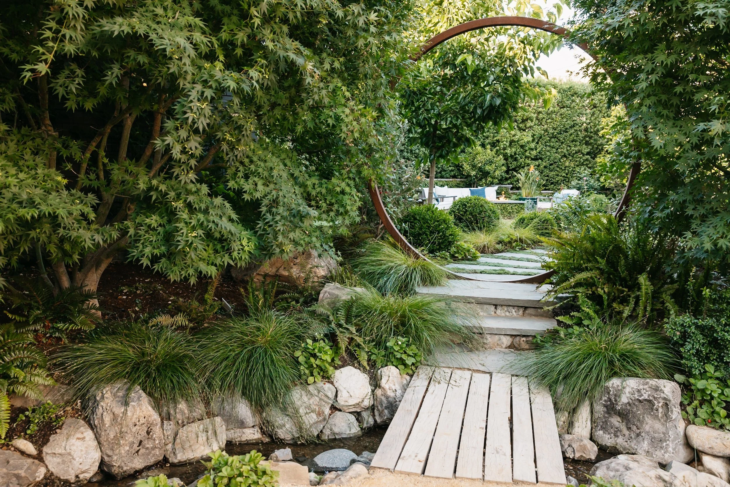 A garden pathway with wooden stairs surrounded by lush green plants and rocks, leading to a sitting area with chairs and pillows in the background.