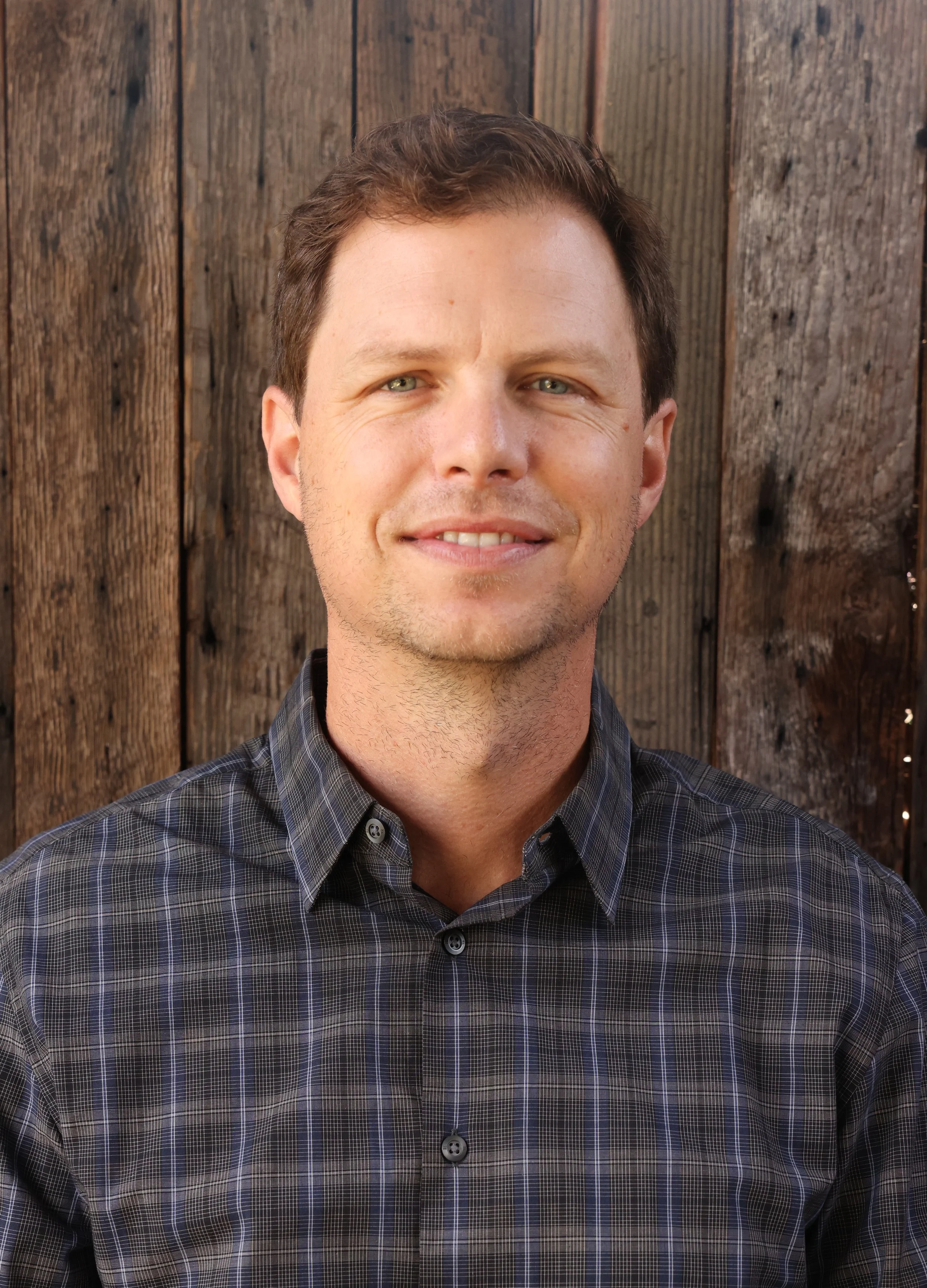 Portrait of a smiling man with brown hair wearing a dark plaid shirt, standing in front of a wooden fence.