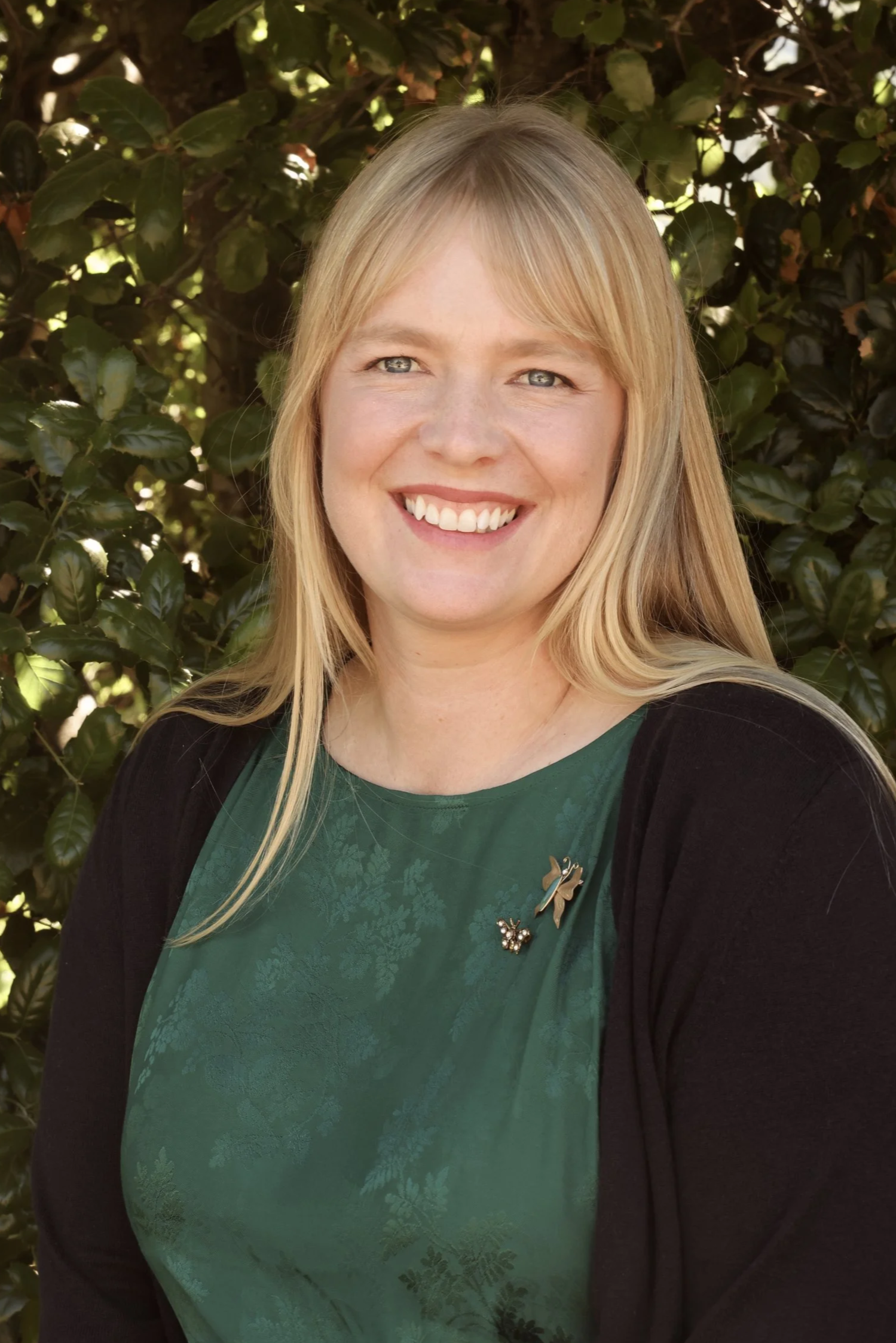 A smiling woman with blonde hair, blue eyes, and light skin, standing outdoors in front of green leafy bushes, wearing a dark cardigan over a green patterned top with decorative pins.