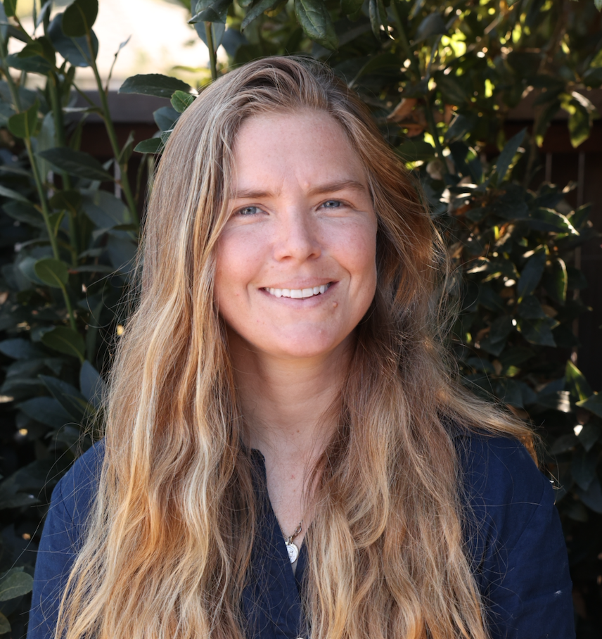 A woman with long, wavy blonde hair smiling outdoors in front of dark green foliage.