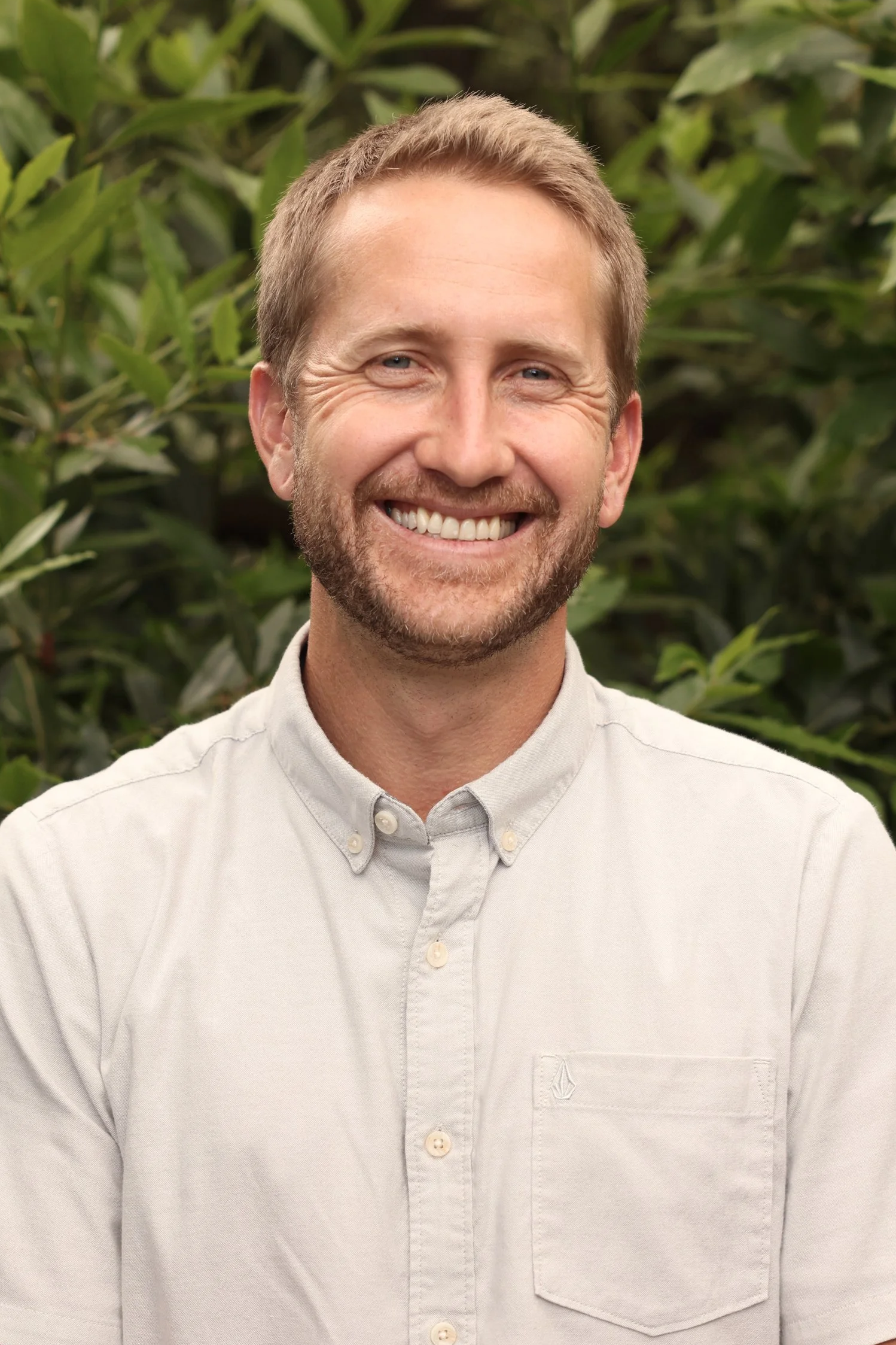 A smiling man with short blond hair and a beard, wearing a light-colored button-up shirt, standing in front of green leafy plants.