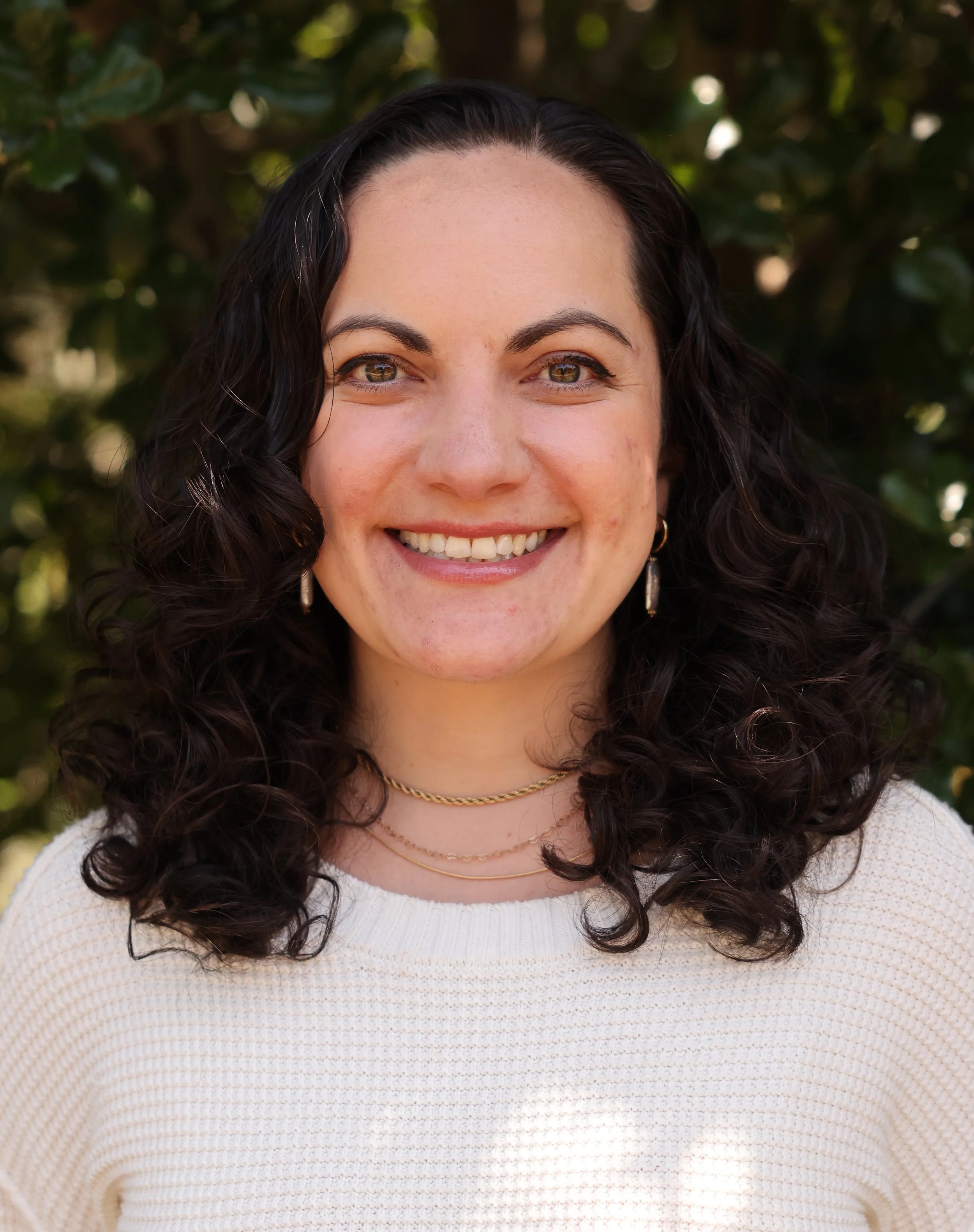 Close-up of a smiling woman with dark, curly hair and green eyes, wearing a white sweater and layered gold necklaces, outdoors with blurred green foliage in the background.