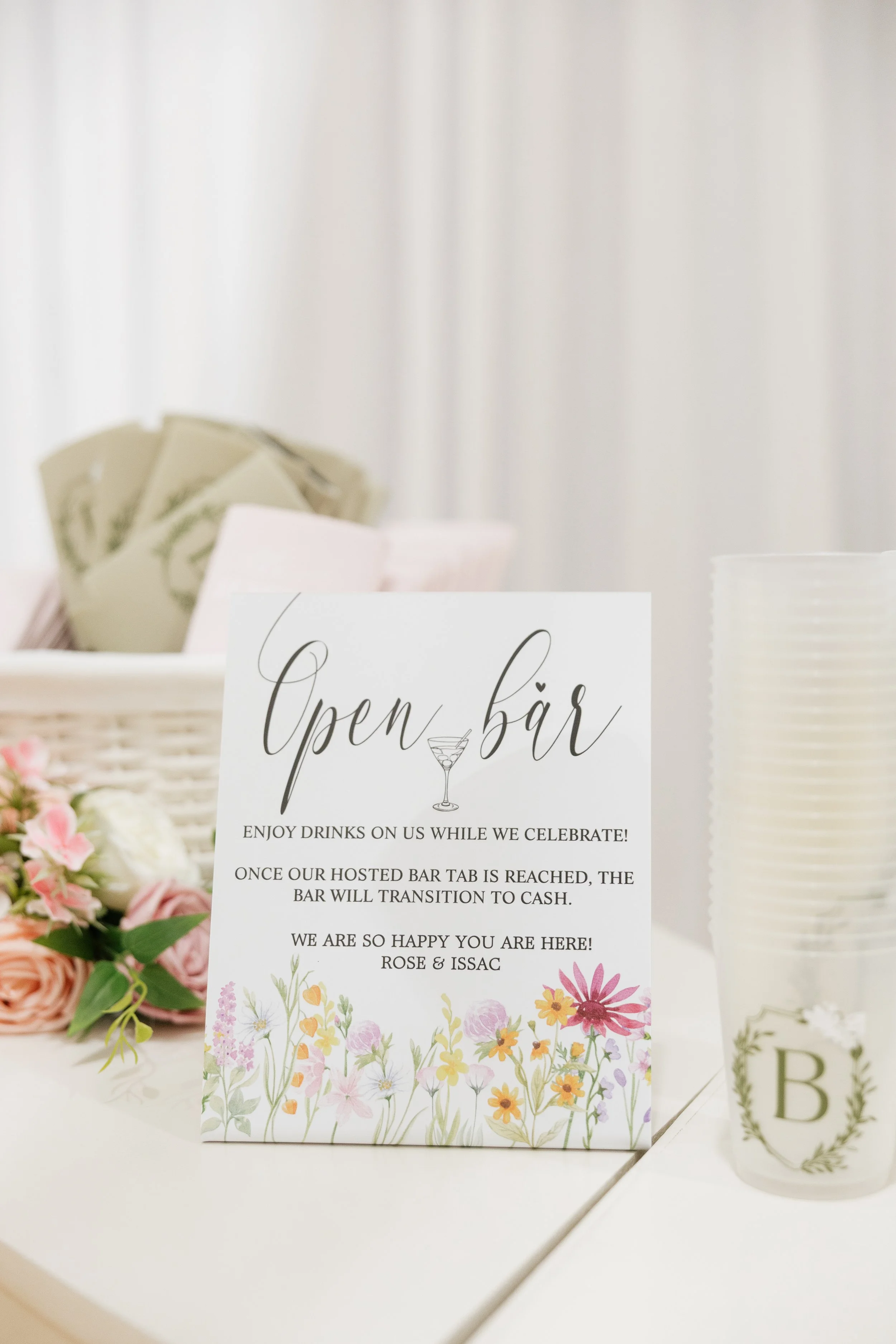 A sign at a wedding reading 'Open bar' with floral illustrations, with a background of flowers.