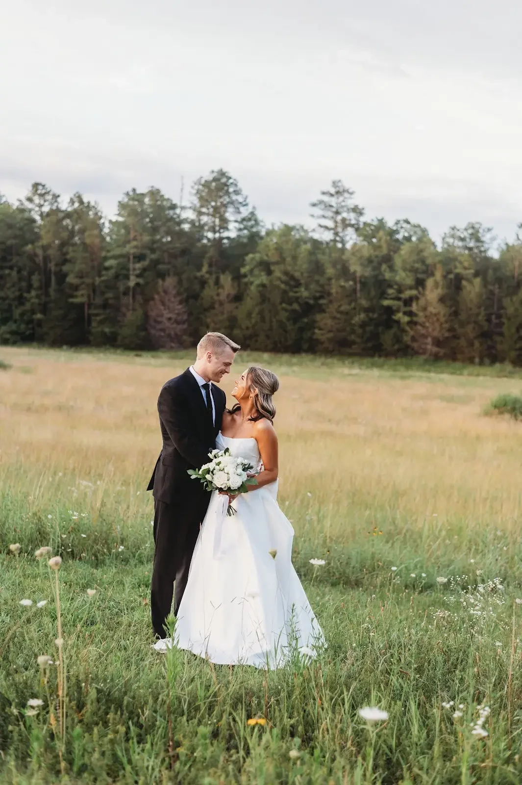 A bride and groom standing in a field, gazing at each other, with trees in the background and the bride holding a bouquet of white flowers.