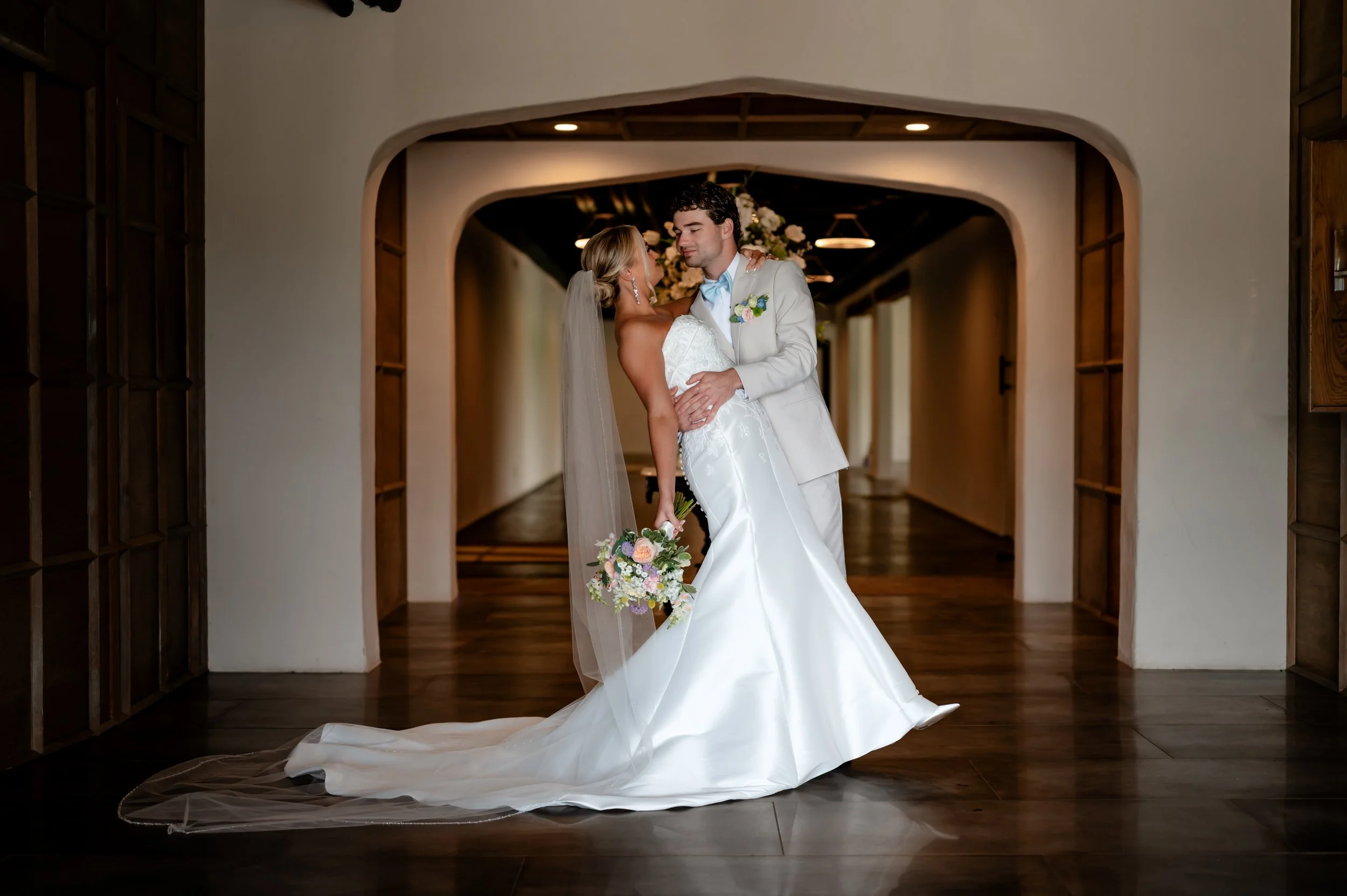 Bride and groom sharing a dance in the entryway during their wedding, with the bride wearing a white wedding gown and veil, holding a bouquet of flowers, and the groom in a light-colored suit, surrounded by a cozy indoor setting with warm lighting.