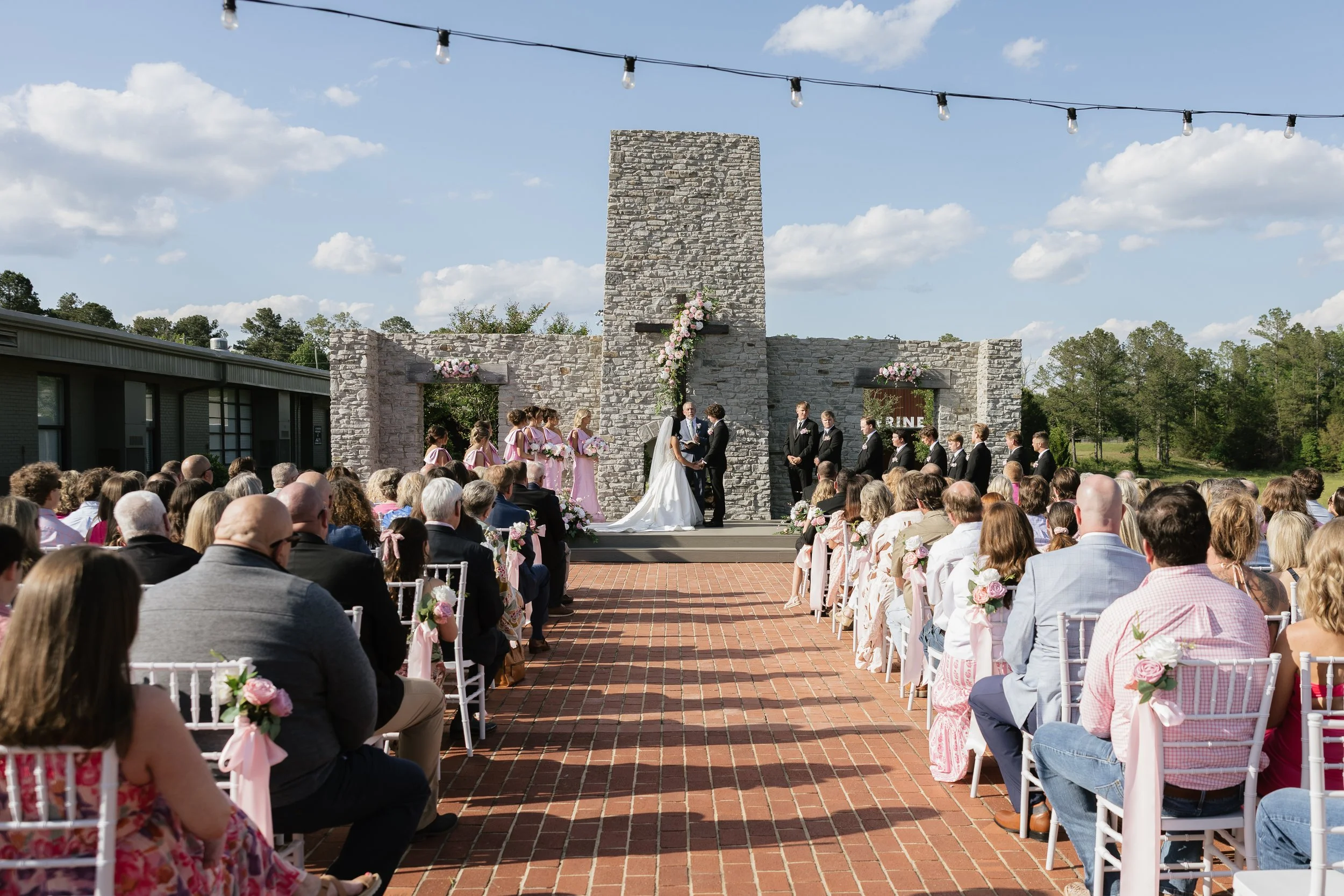 A large outdoor wedding ceremony with guests seated facing an altar made of stone with a cross and flowers. The bride and groom are exchanging vows underneath a stone tower with floral decorations. String lights hang above the ceremony area on a clear day with a blue sky and some clouds.
