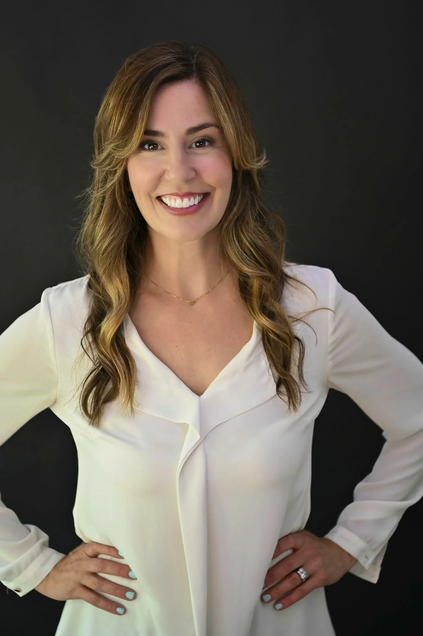 A woman with long wavy light brown hair and a wide smile, wearing a white blouse, standing against a dark background.