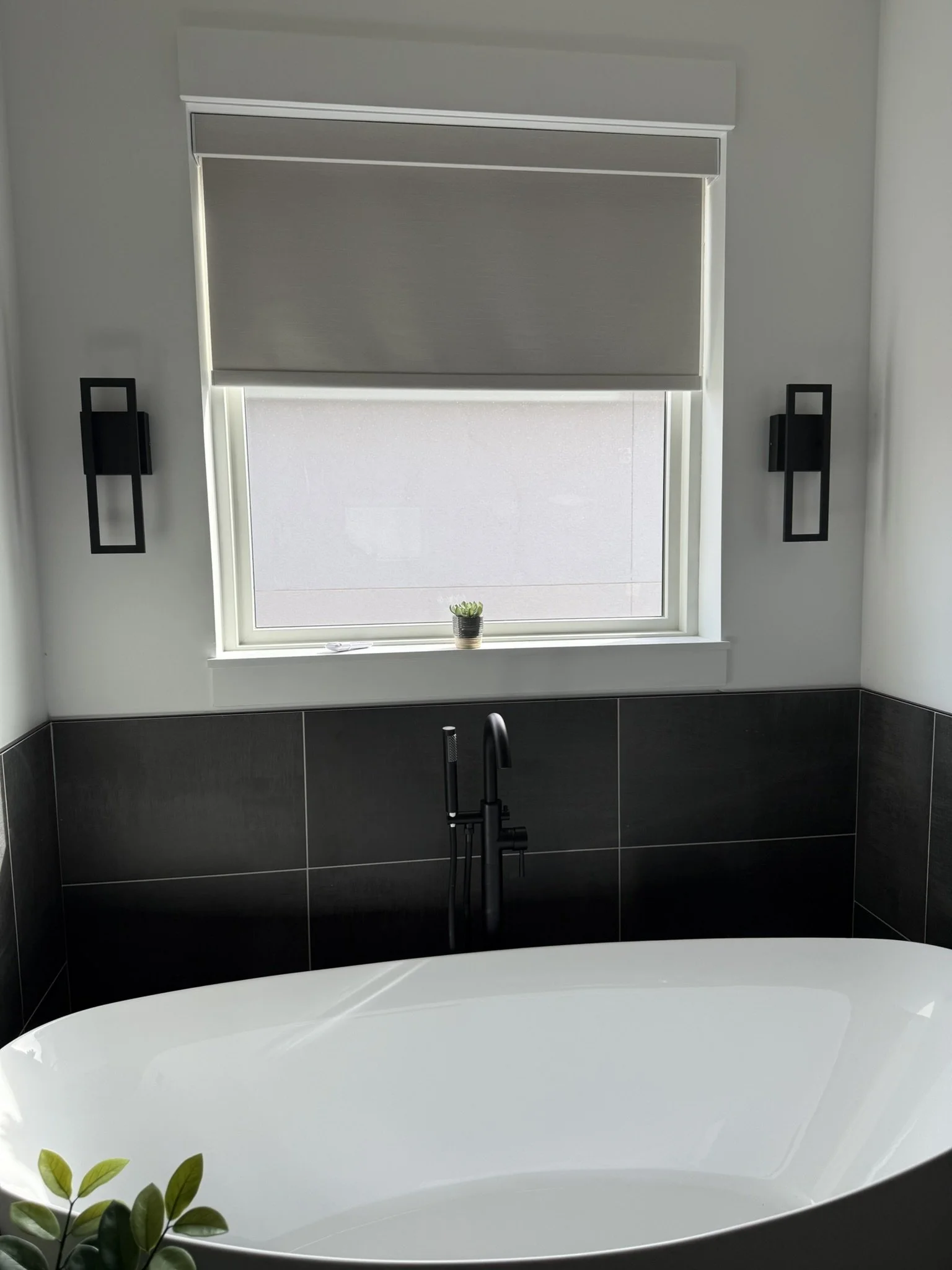 Modern bathroom with a freestanding bathtub, black faucet, black wall-mounted lights, a window with a blind, and a small potted plant on the windowsill.