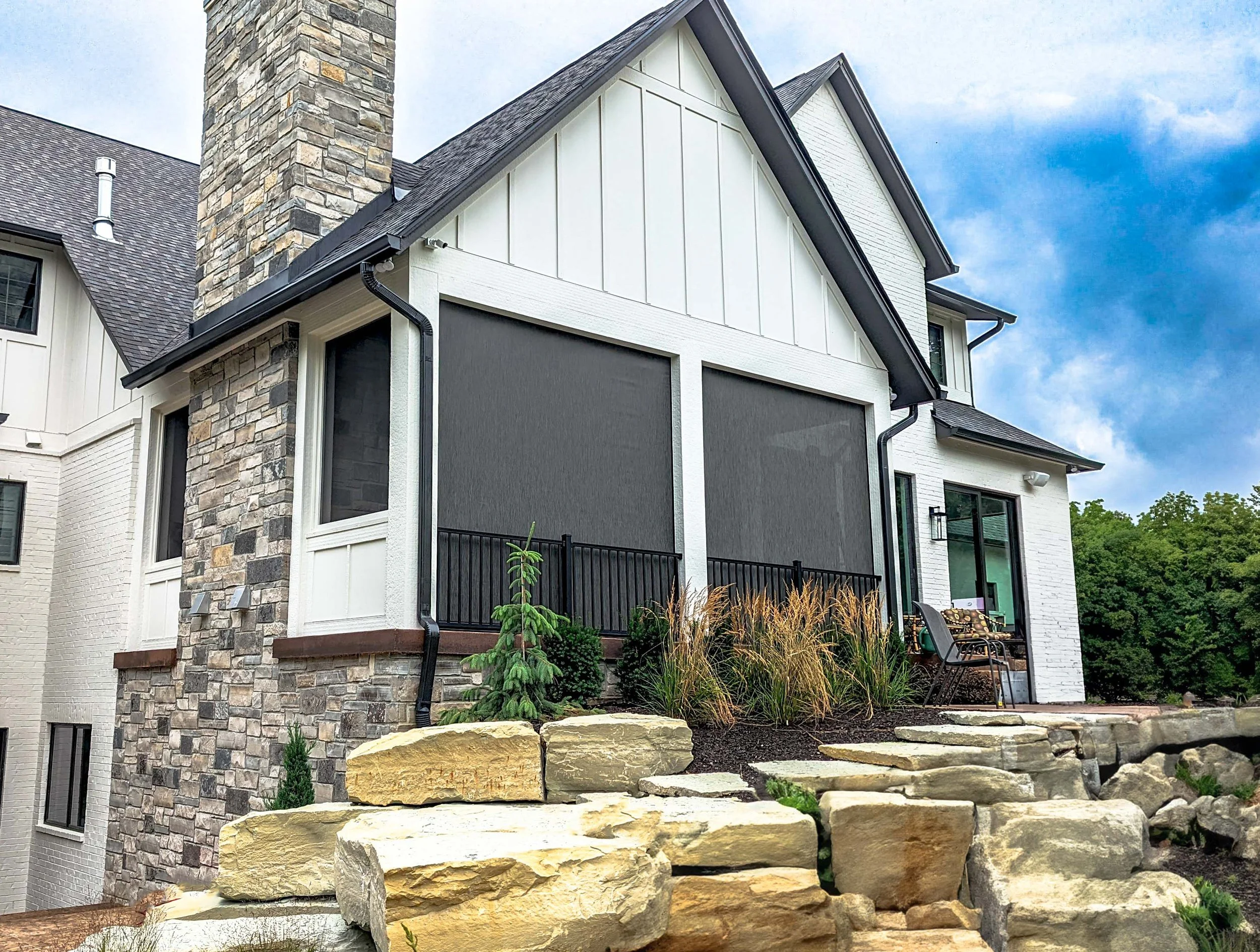 A modern house with a stone chimney, dark gray garage door, and white siding. The house has a sloped roof and a small landscaped yard with plants, rocks, and a stone pathway.