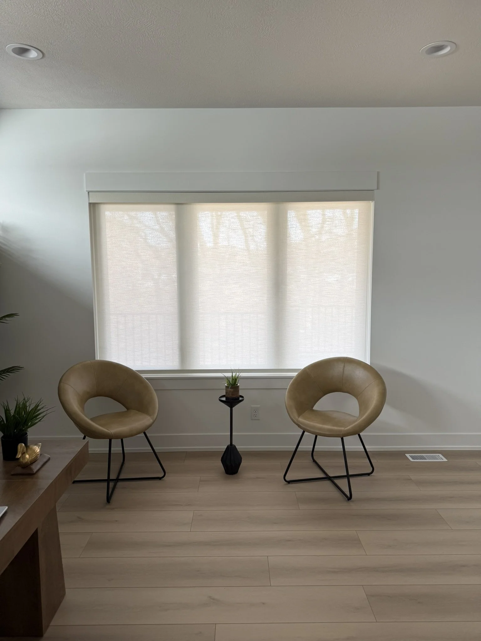 A cozy living space with a large window covered by beige blinds, two beige modern chairs with black metal legs, a small black side table with a plant, and wooden flooring.