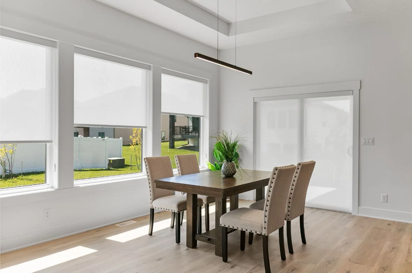 A bright dining room with large windows, a wooden table, beige upholstered chairs, and a potted plant in the center.