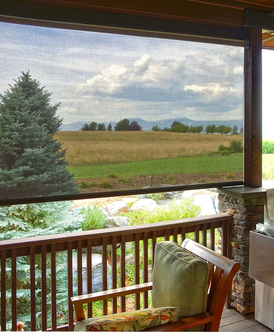 A view of a grassy field, trees, and mountains in the distance seen from a porch with a wooden railing, a cushioned chair, and a grill.