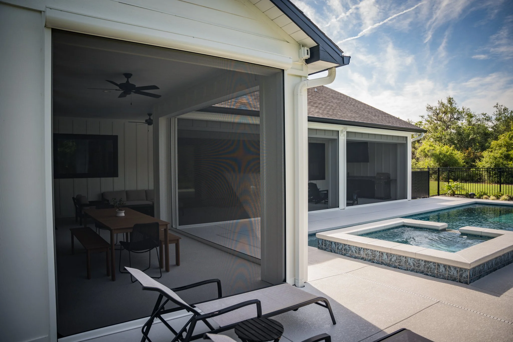 A backyard area with a swimming pool, hot tub, safety fence, and a covered patio with a ceiling fan and indoor furniture visible through sliding glass doors.