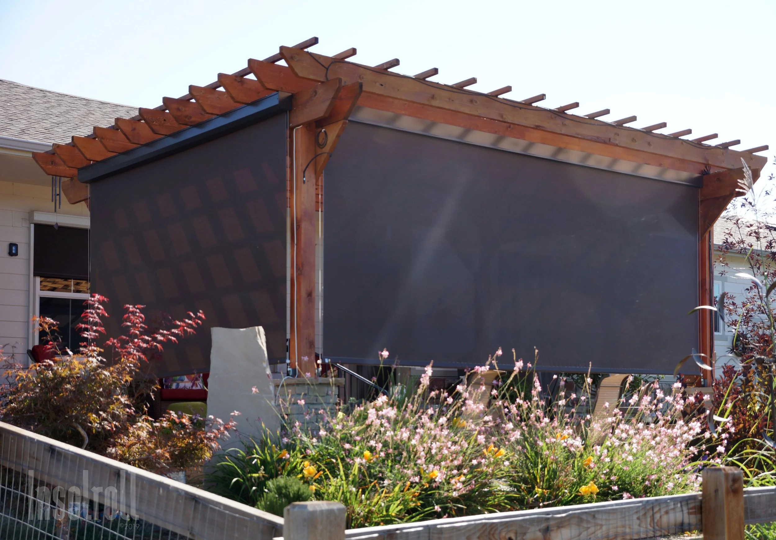 A garden bed with flowering plants in front of a black shade cloth and wooden pergola structure with unfinished roof framing, in a residential backyard.