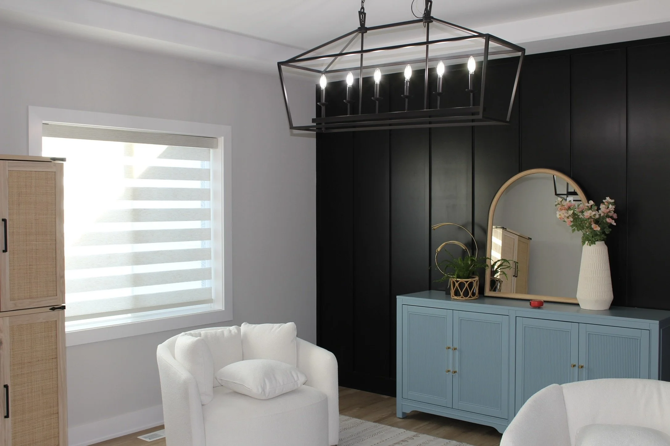 Interior of a living room with a black wall, a window with white shades, a white armchair, a light blue cabinet with gold handles, a large mirror, a vase with pink flowers, and a modern black chandelier with six candle-shaped lights.