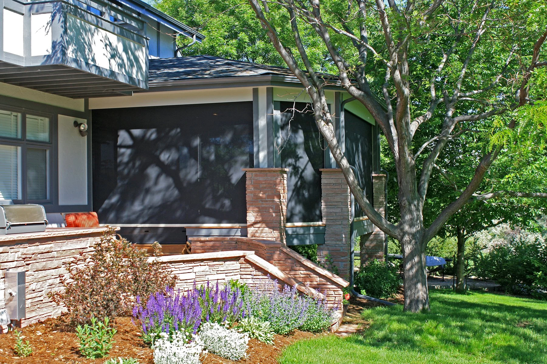 A house with a garden, featuring a large tree and colorful flowers, a brick staircase, and a screened porch with windows.