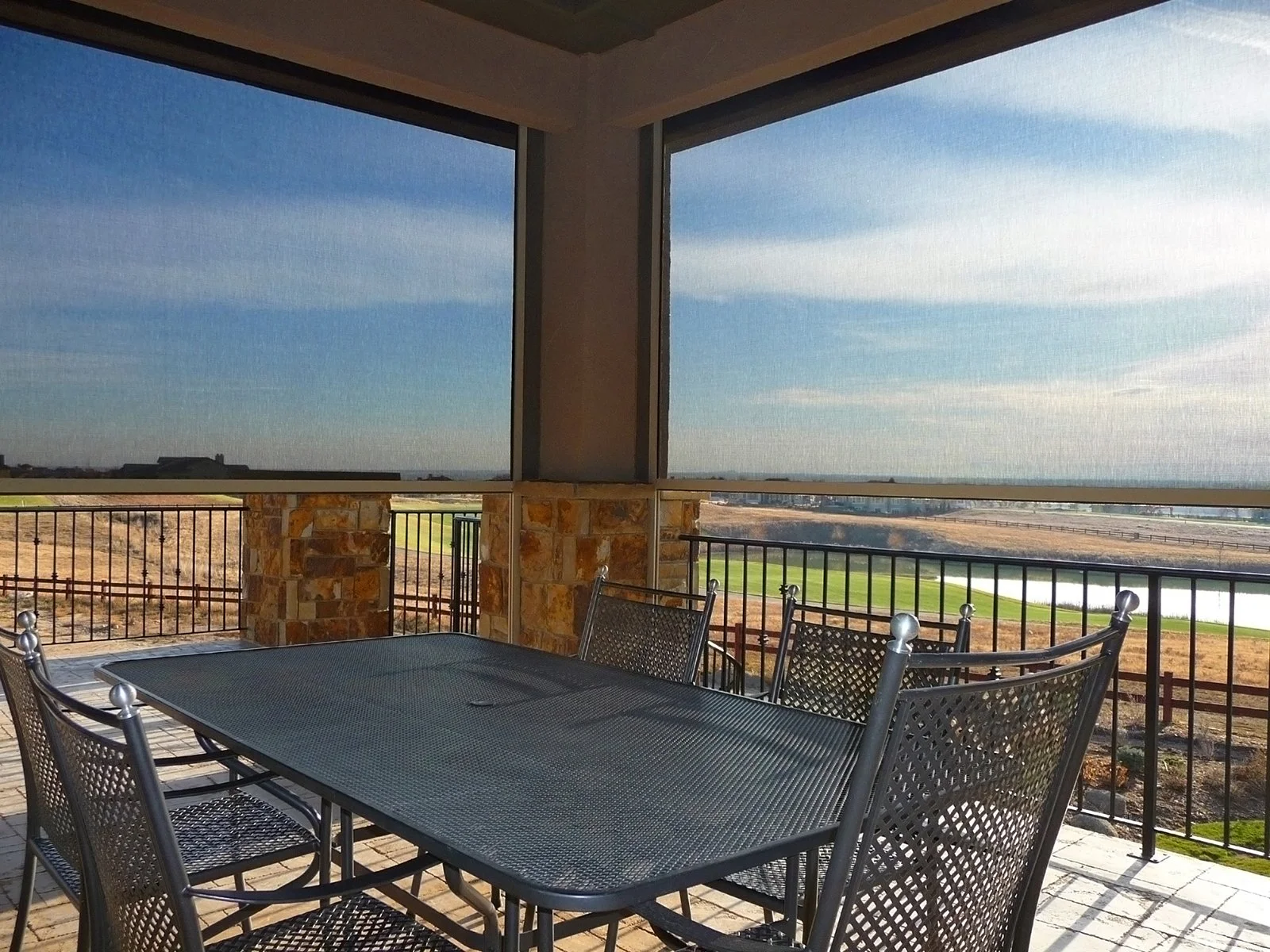 Covered patio with black metal dining table and chairs overlooking a golf course with water hazards and a blue sky with scattered clouds.