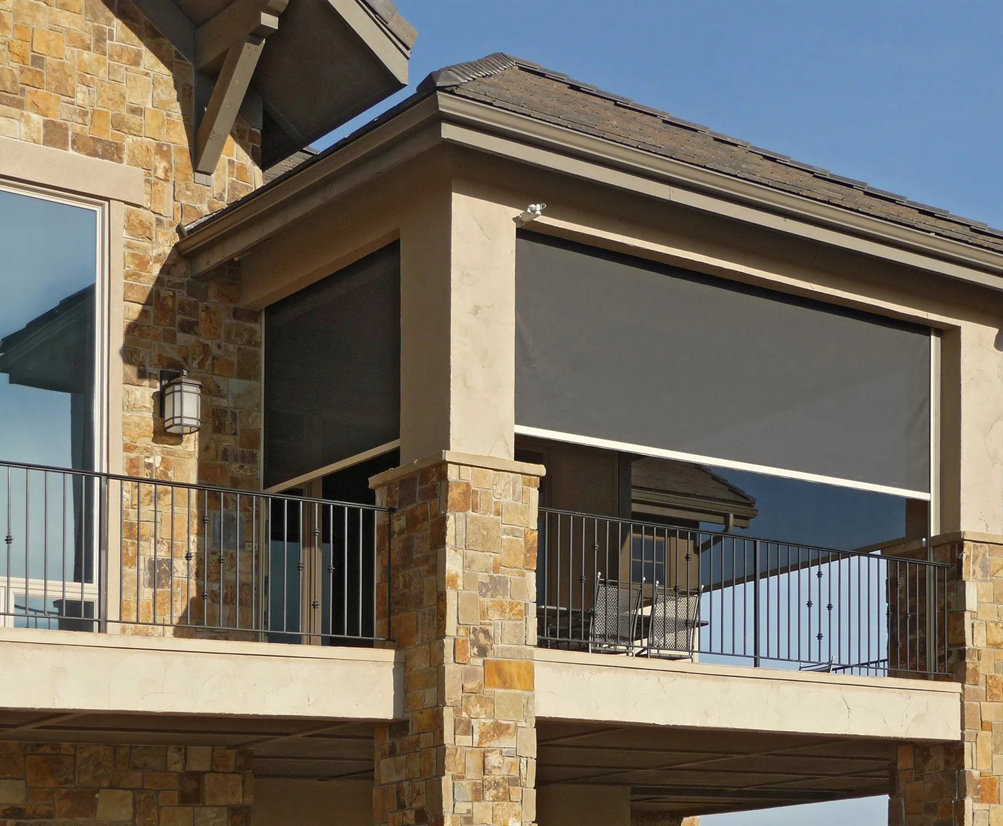 Close-up of a building balcony with brick pillars, glass windows, metal railing, and a roll-up shade, against a blue sky background.