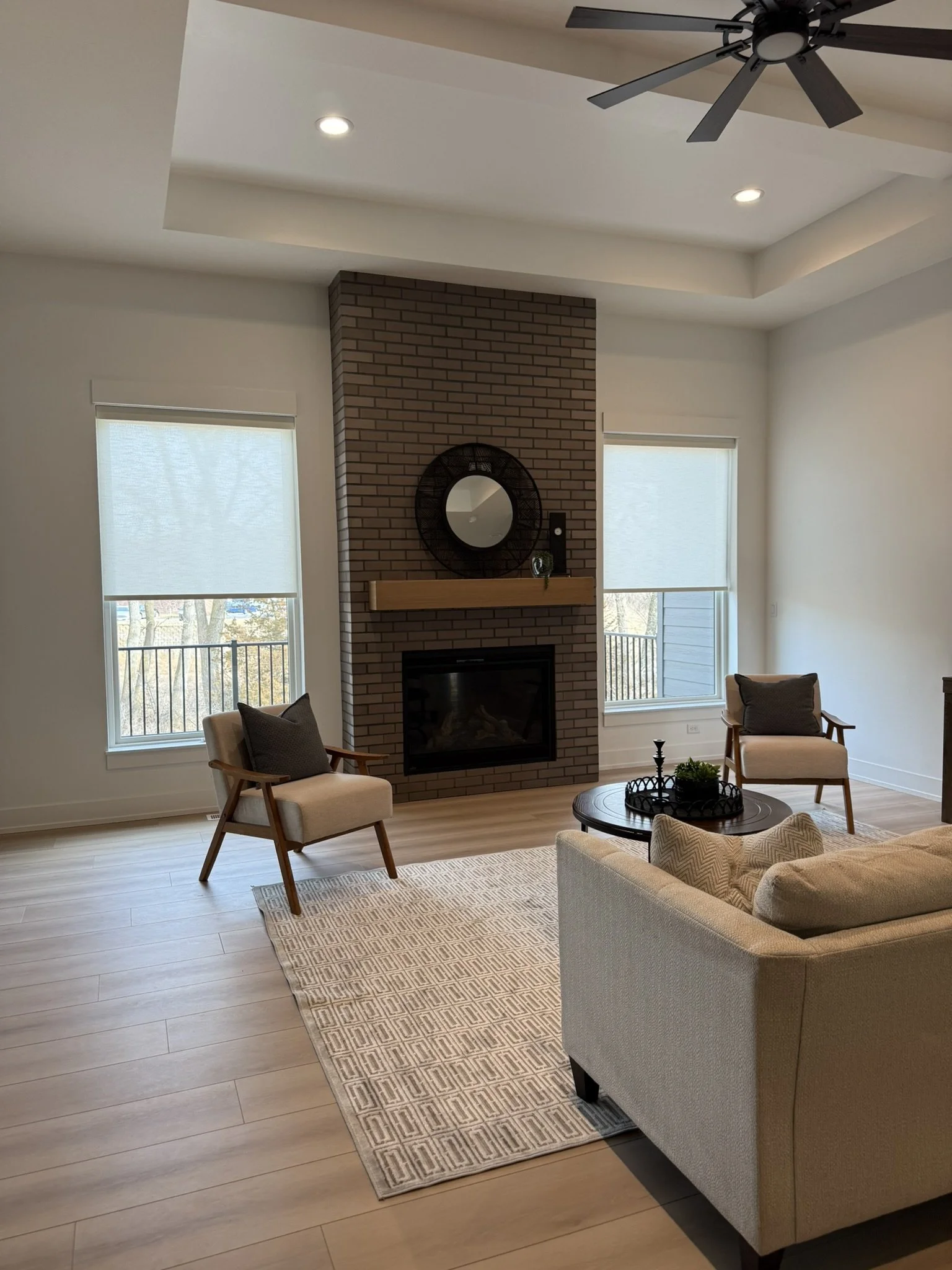 Living room with a brick fireplace, two windows with white blinds, armchairs, a coffee table, a beige sofa, ceiling fan, and hardwood floors.