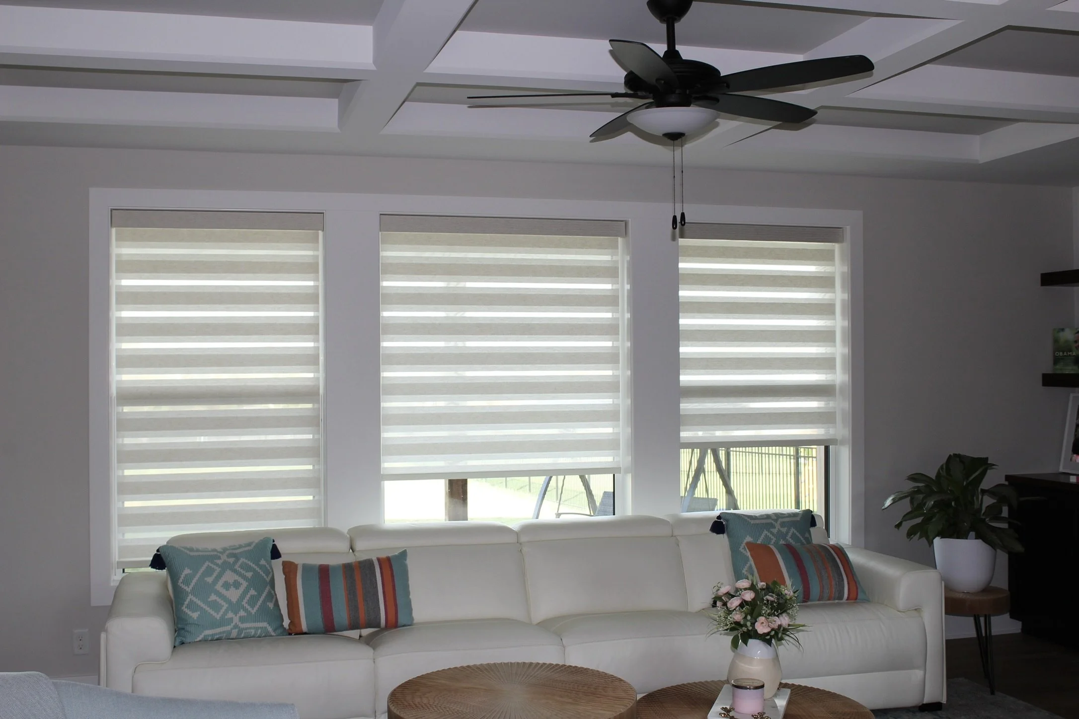 Living room with three large windows covered with beige zebra shades, white walls, ceiling fan with light, white sofa with colorful patterned pillows, a round rattan coffee table, a small flower arrangement, and a small side table with a candle.