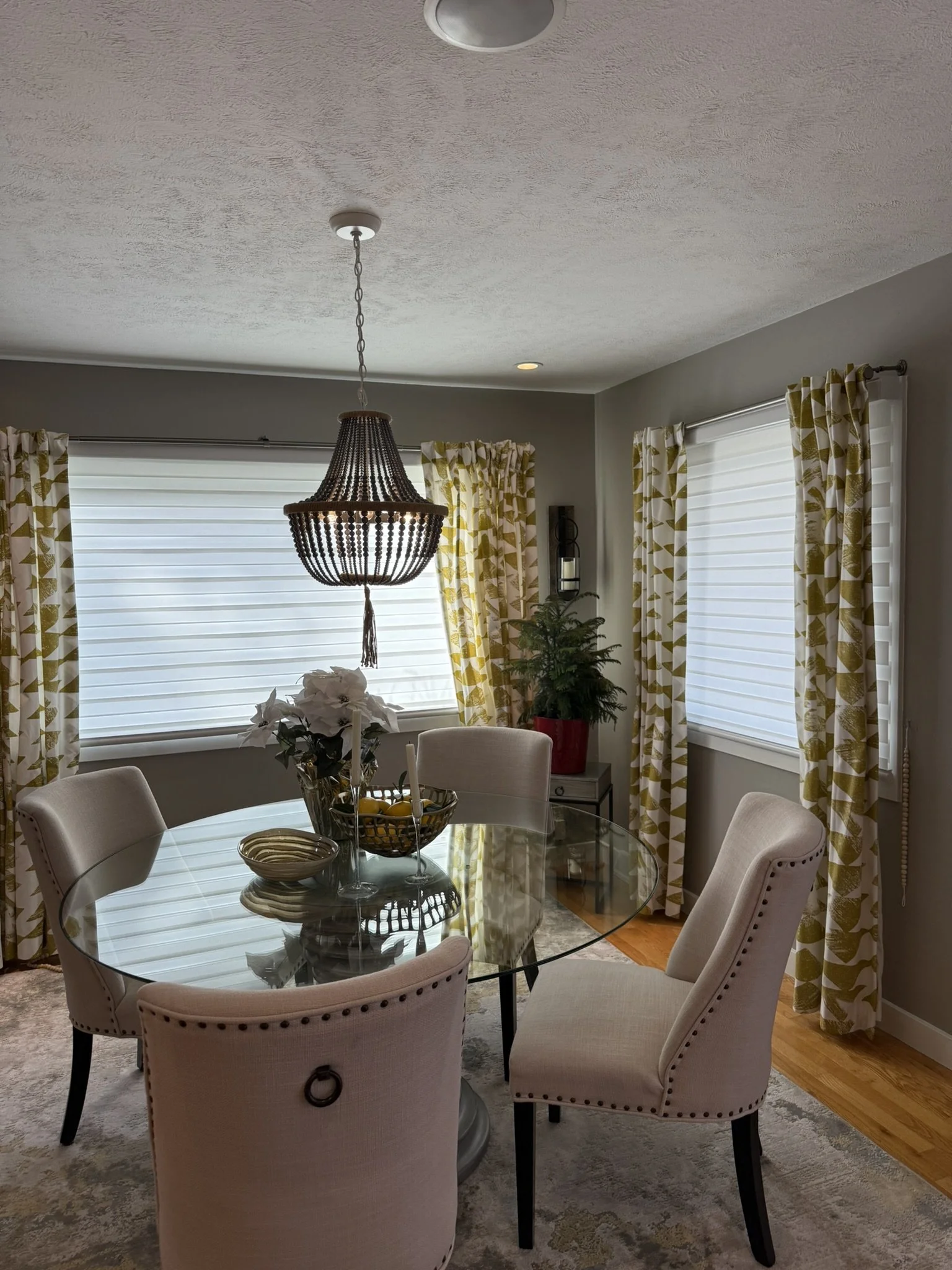 A living room with a round glass dining table surrounded by beige upholstered chairs, a chandelier hanging from the ceiling, large windows with white blinds, yellow and white patterned curtains, a small Christmas tree, and a potted plant.