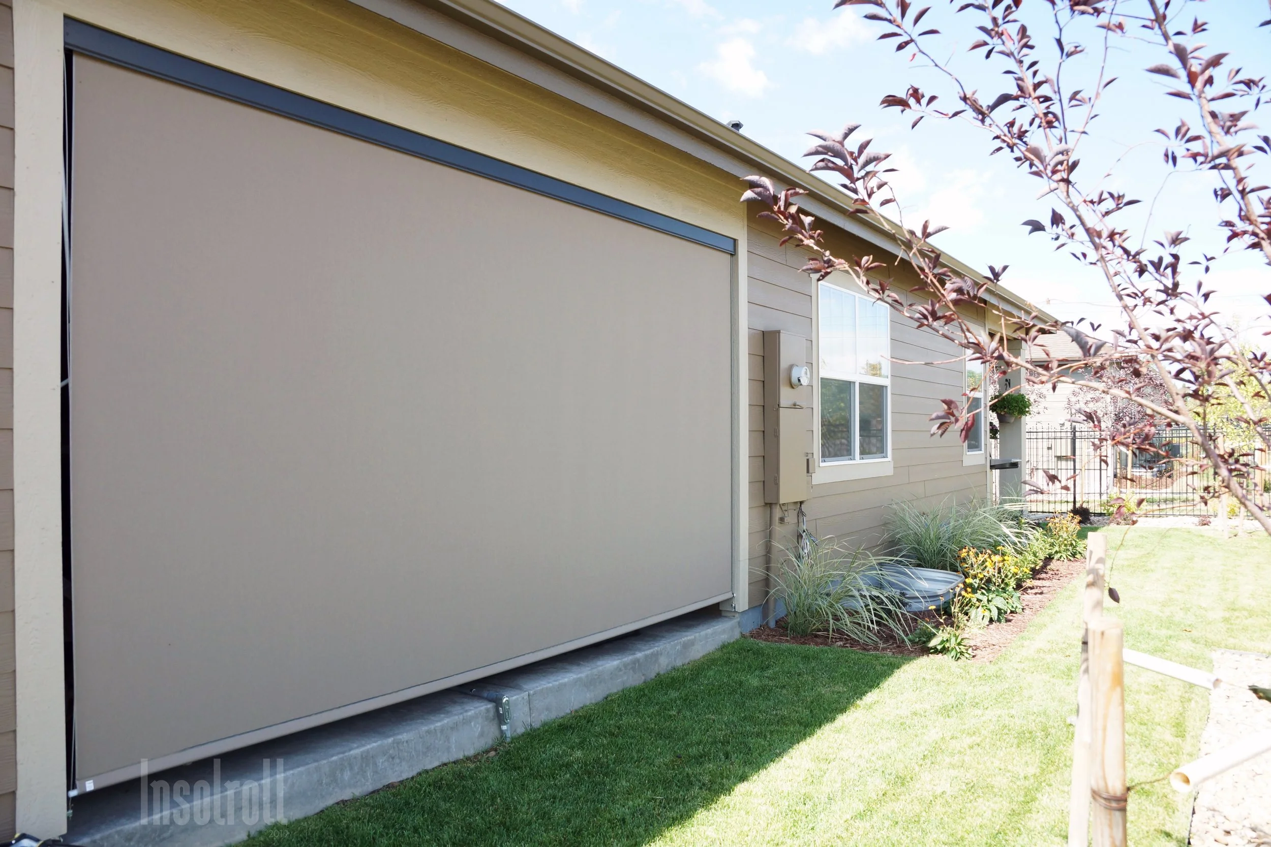 A modern house with a beige exterior and a large beige garage door, surrounded by a well-maintained garden with small trees, bushes, and a green lawn. A black metal fence is visible in the background, and the sky is partly cloudy.