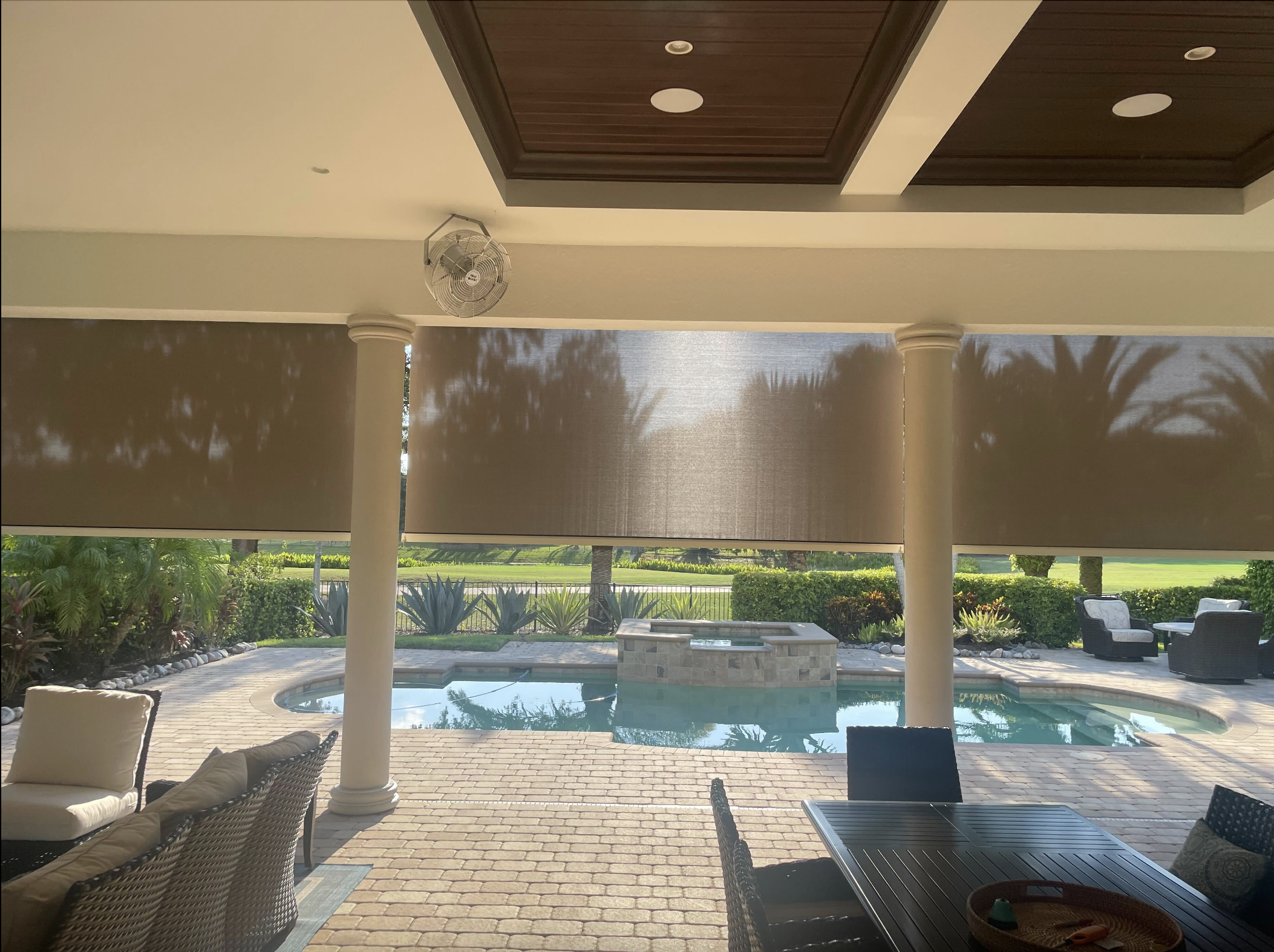 A backyard view of a patio area with wicker chairs, a dining table, and a pool, shaded by a roof with wooden panels, overlooking a well-maintained garden and palm trees.