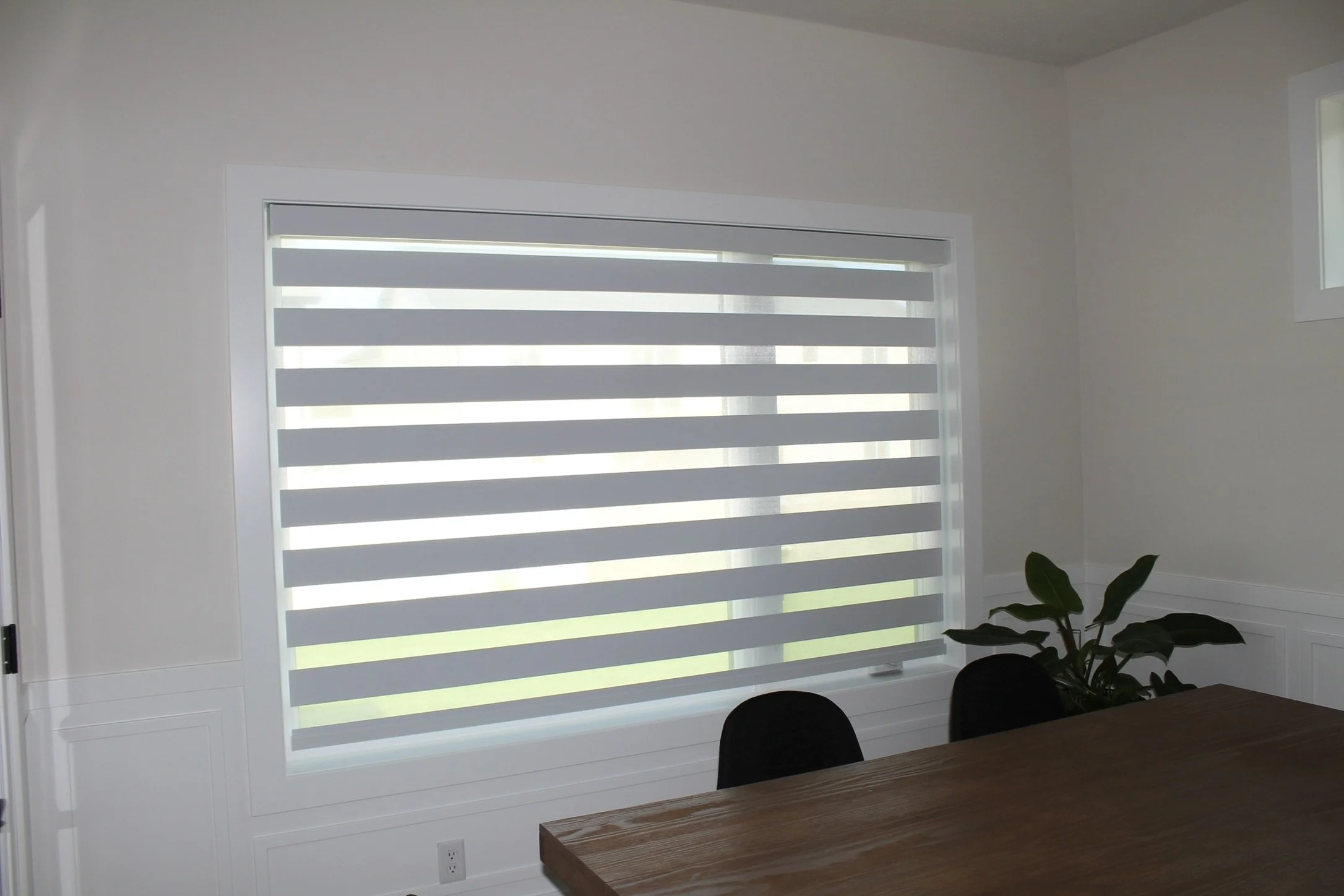 A room with a large window featuring horizontal zebra blinds, a white wall, a wooden table, black chairs, and a green potted plant.