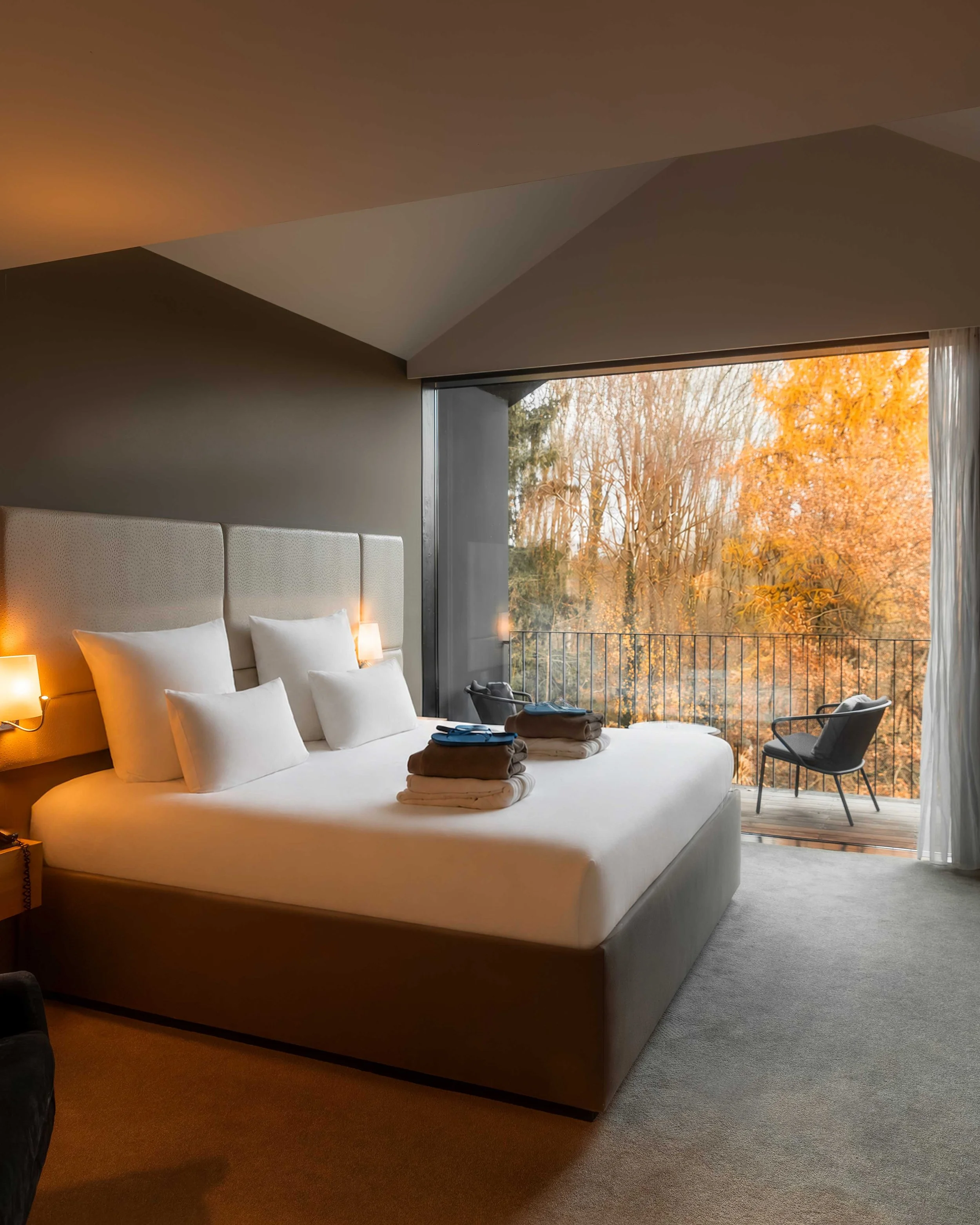 Chambre avec un lit blanc, oreillers et serviettes pliées, vue sur une fenêtre avec des arbres aux feuillages orange.