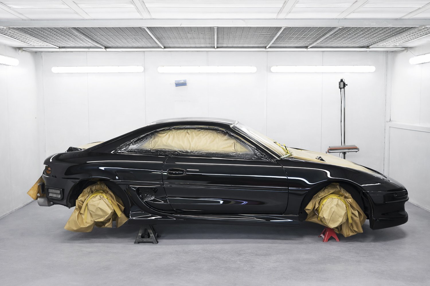 Black sports car in a professional paint booth with masking paper on windows and wheels, elevated on jack stands.