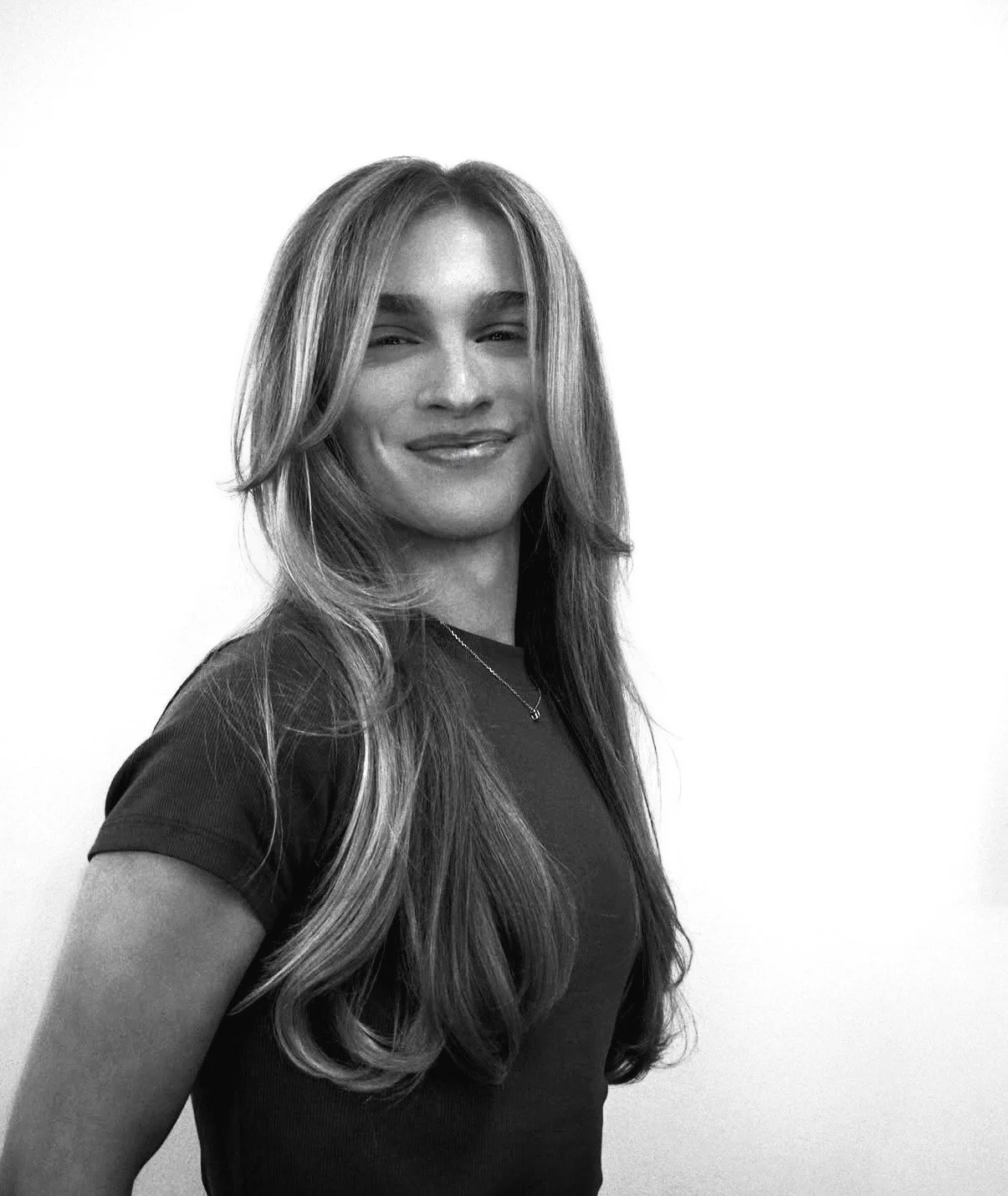 Black and white portrait of a young woman with long wavy hair, wearing a sleeveless top, layered necklaces, and bracelets, looking confidently at the camera with a slight smile.