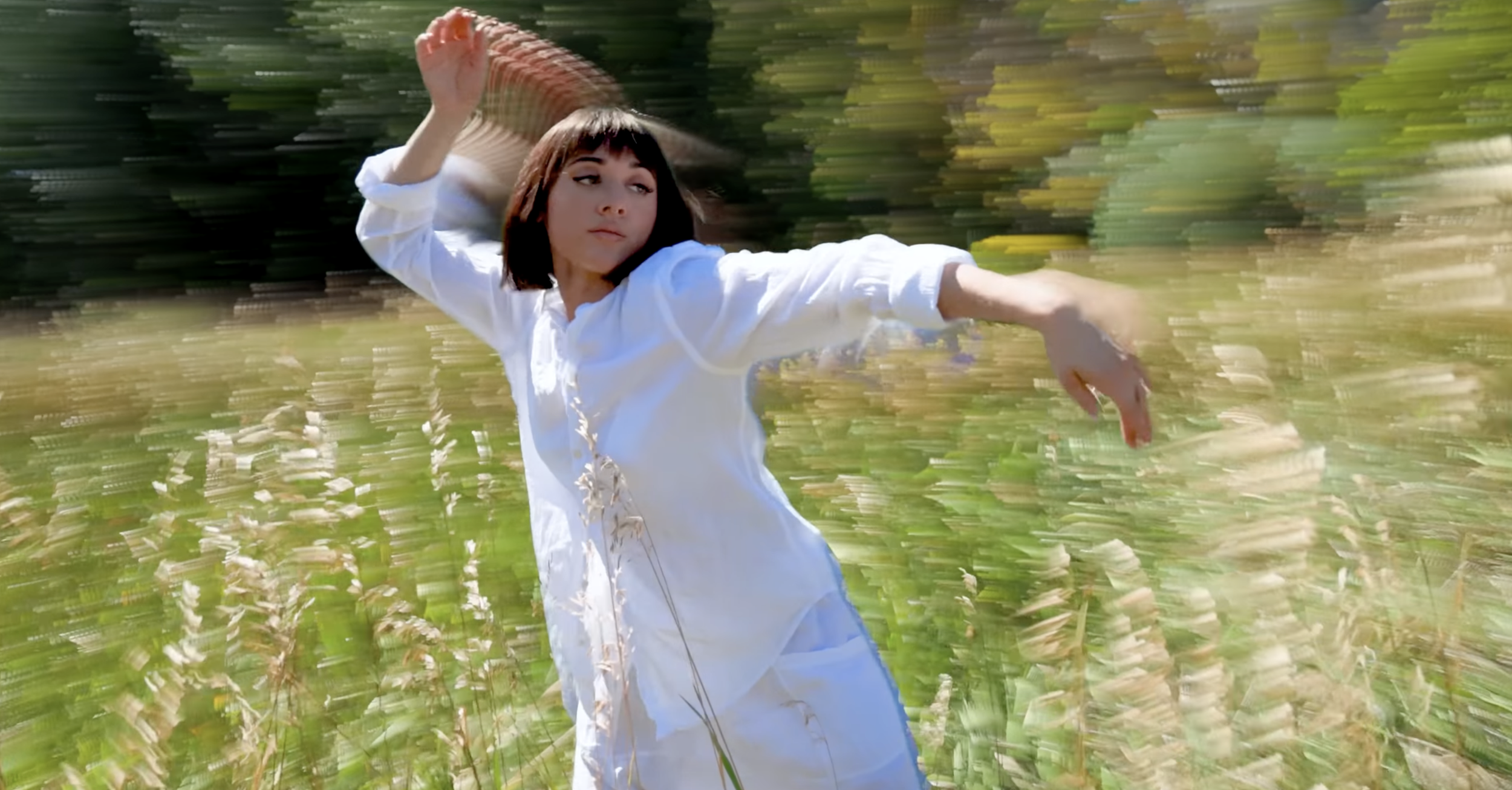 A woman with short dark hair in a white dress holding a fan behind her head while standing in a green outdoor setting, with a blurred background.