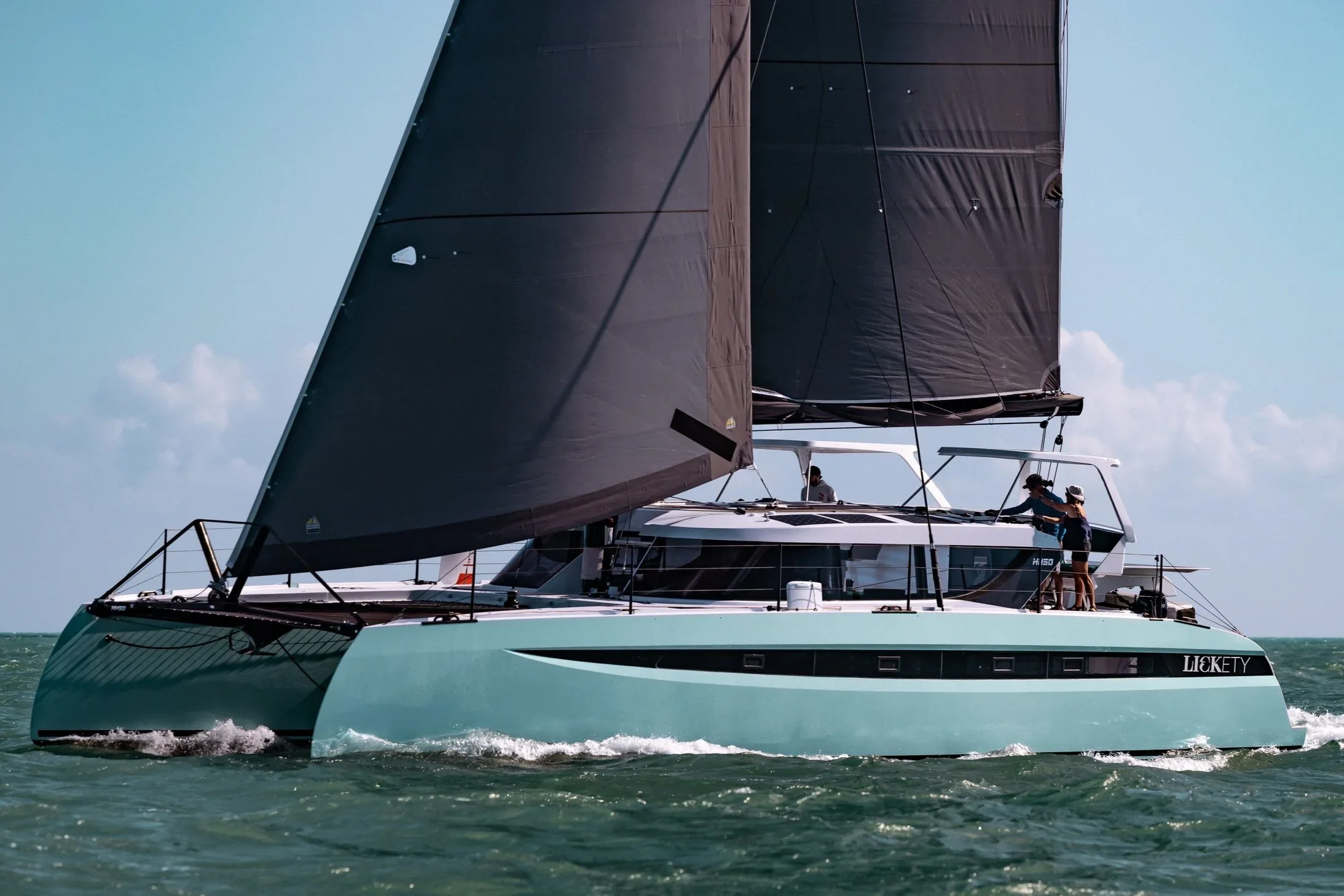 A yacht with large black sails sailing on the ocean with a clear blue sky in the background. Two people are visible on the deck, one standing near the railing and the other sitting or leaning against a surface.