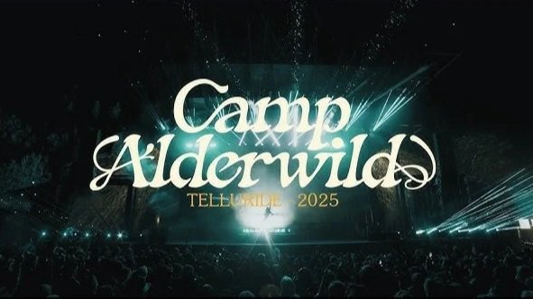 Event stage with large screens displaying 'Camp Alderwild Tell Tale 2025' at night, surrounded by audience and stage lights.