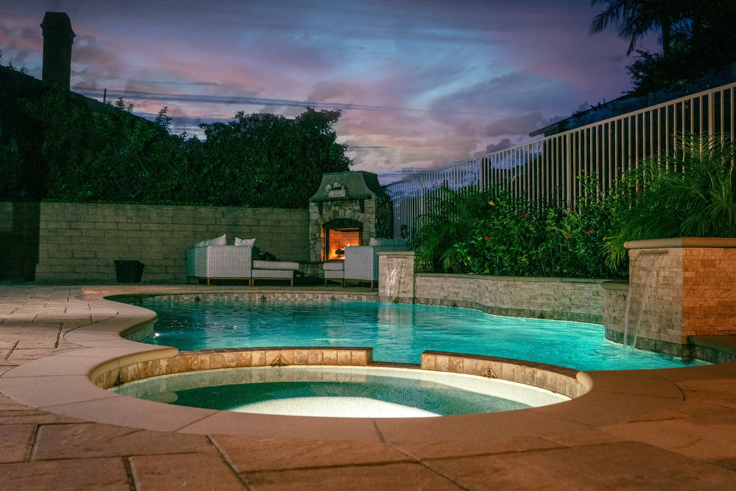 Quiet backyard swimming pool with a hot tub, outdoor sofa set, fireplace, and a colorful evening sky.