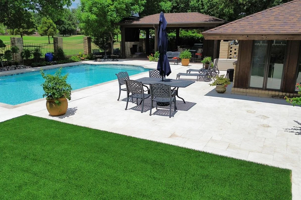 Beautiful backyard featuring a swimming pool with clear blue water, surrounded by a white stone patio. There is a dining table with six chairs and a closed umbrella in the center. Several potted plants are placed around the patio. A covered outdoor seating area with furniture is visible in the background, along with lush green trees and a fenced lawn.