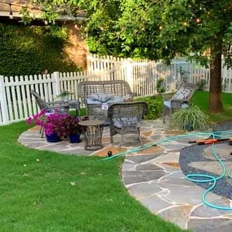 Outdoor patio area with a black metal table and chairs, surrounded by a white picket fence, lush green grass, colorful flowers, and trees.