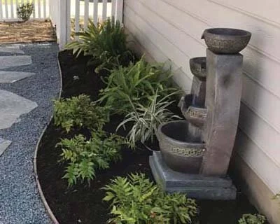 A garden with various green plants next to a gray stone water fountain against a white house wall.
