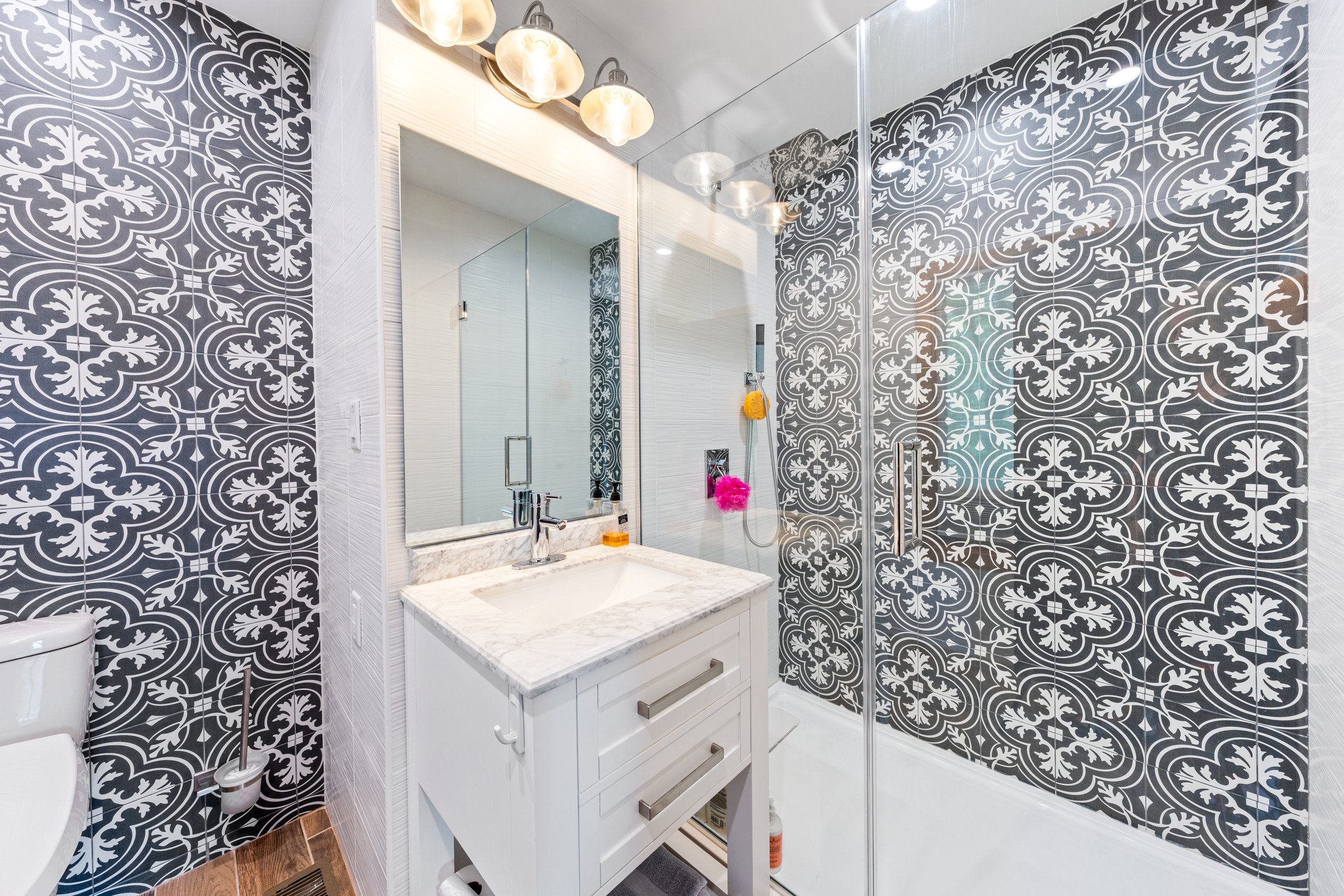 Bathroom with black and white patterned tiles on the walls, a white vanity with a marble countertop, a mirrored medicine cabinet, and a glass-enclosed shower with colorful bath poufs hanging inside.